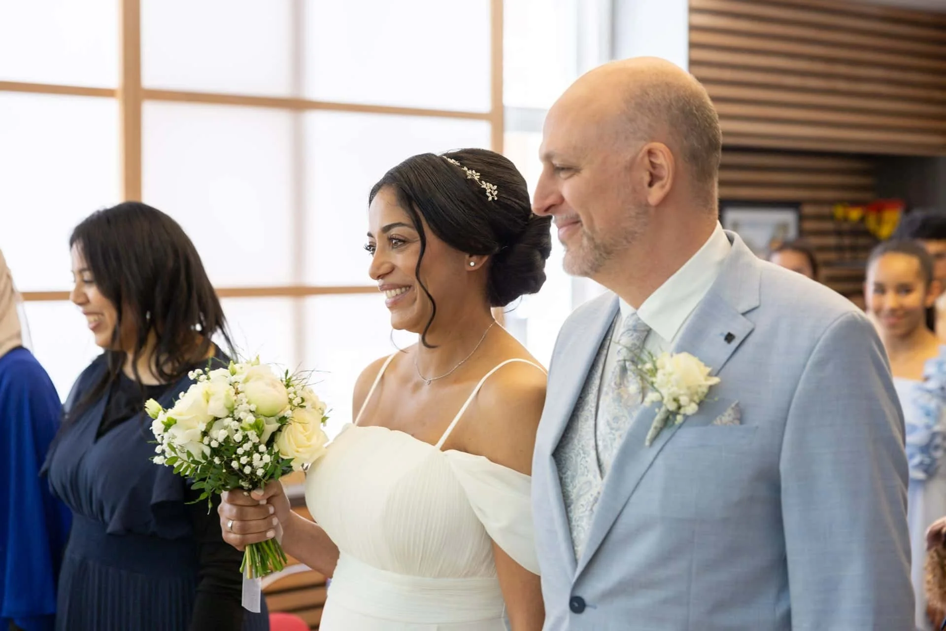 Un couple de mariés lors de la cérémonie, sourire, femme tenant un bouquet de fleurs, homme en costume clair, entourés d'invités heureux.