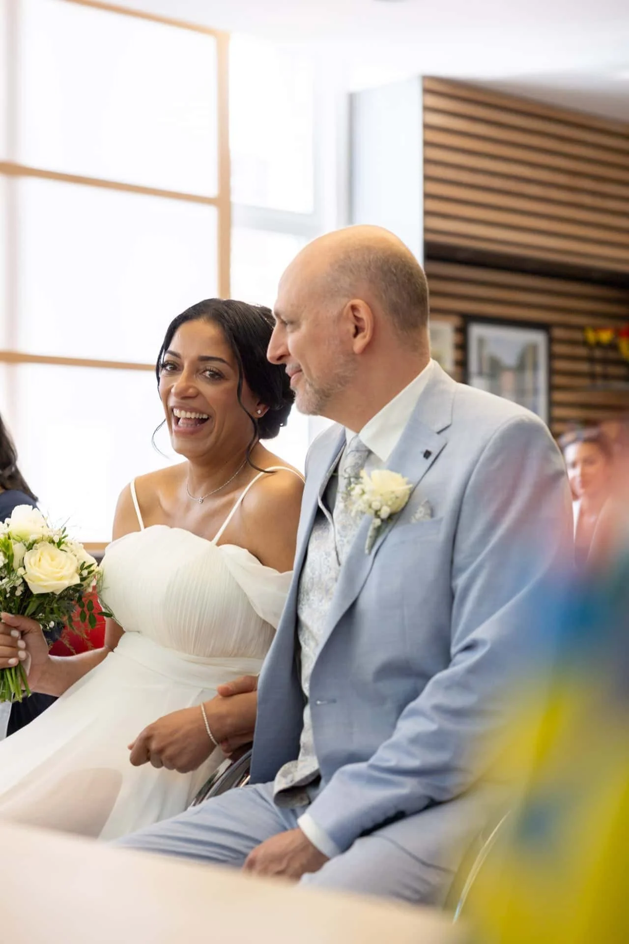Un couple lors d'un mariage, la femme en robe blanche souriante et l'homme en costume gris, assis ensemble dans une salle de cérémonie.