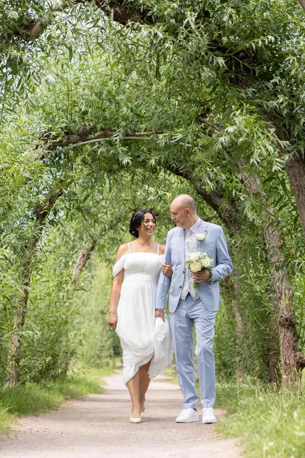 Un couple en mariage marchant dans un chemin bordé d'arbres verts.