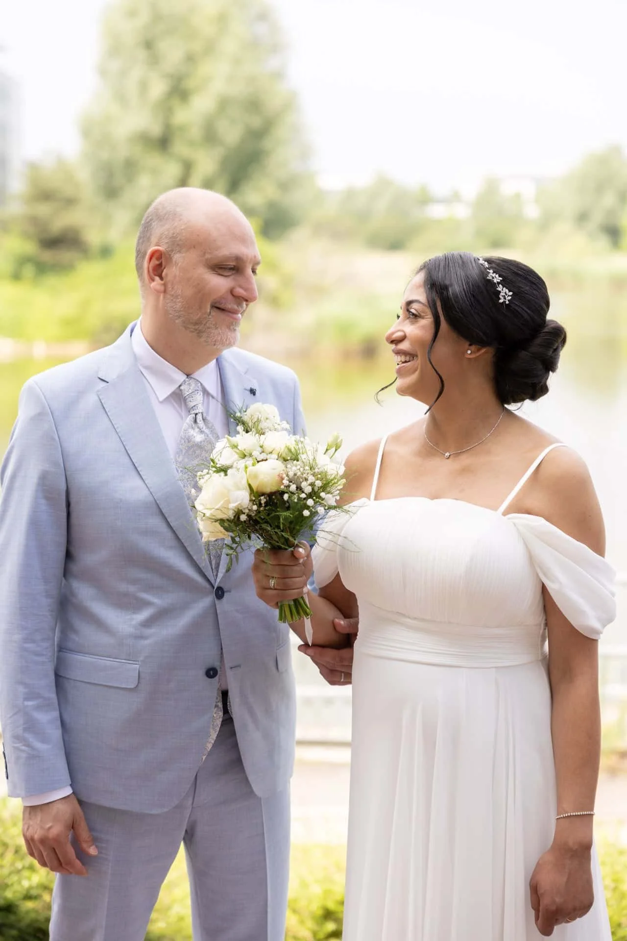 Un couple de mariés, un homme en costume gris clair et une femme en robe blanche, s'échangeant un sourire en se tenant la main lors d'un mariage en extérieur.