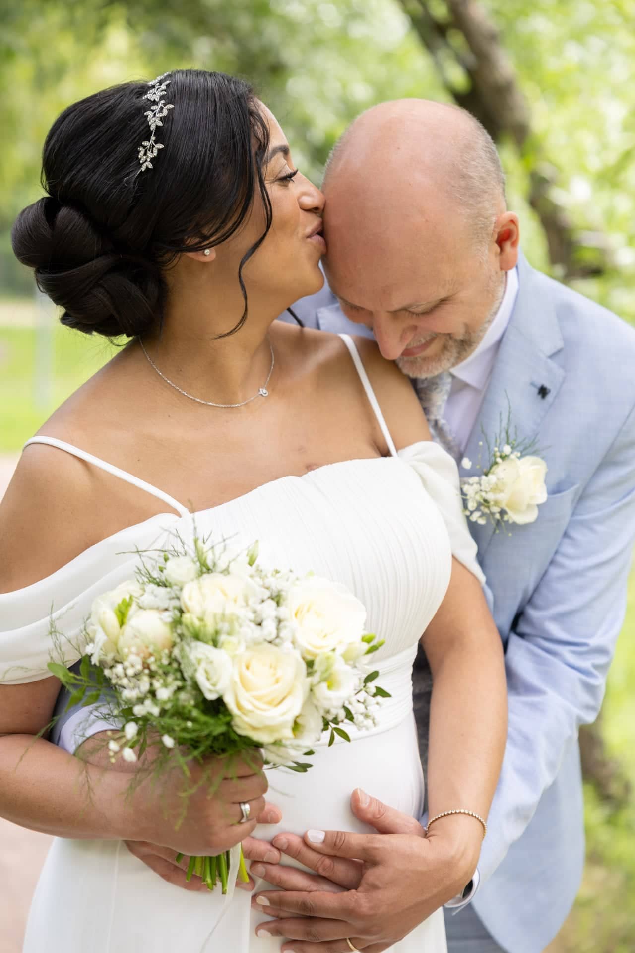 Un couple de mariés partage un moment tendre en plein air, elle en mauve et lui en gris clair, avec un bouquet de fleurs blanches, entouré de verdure.
