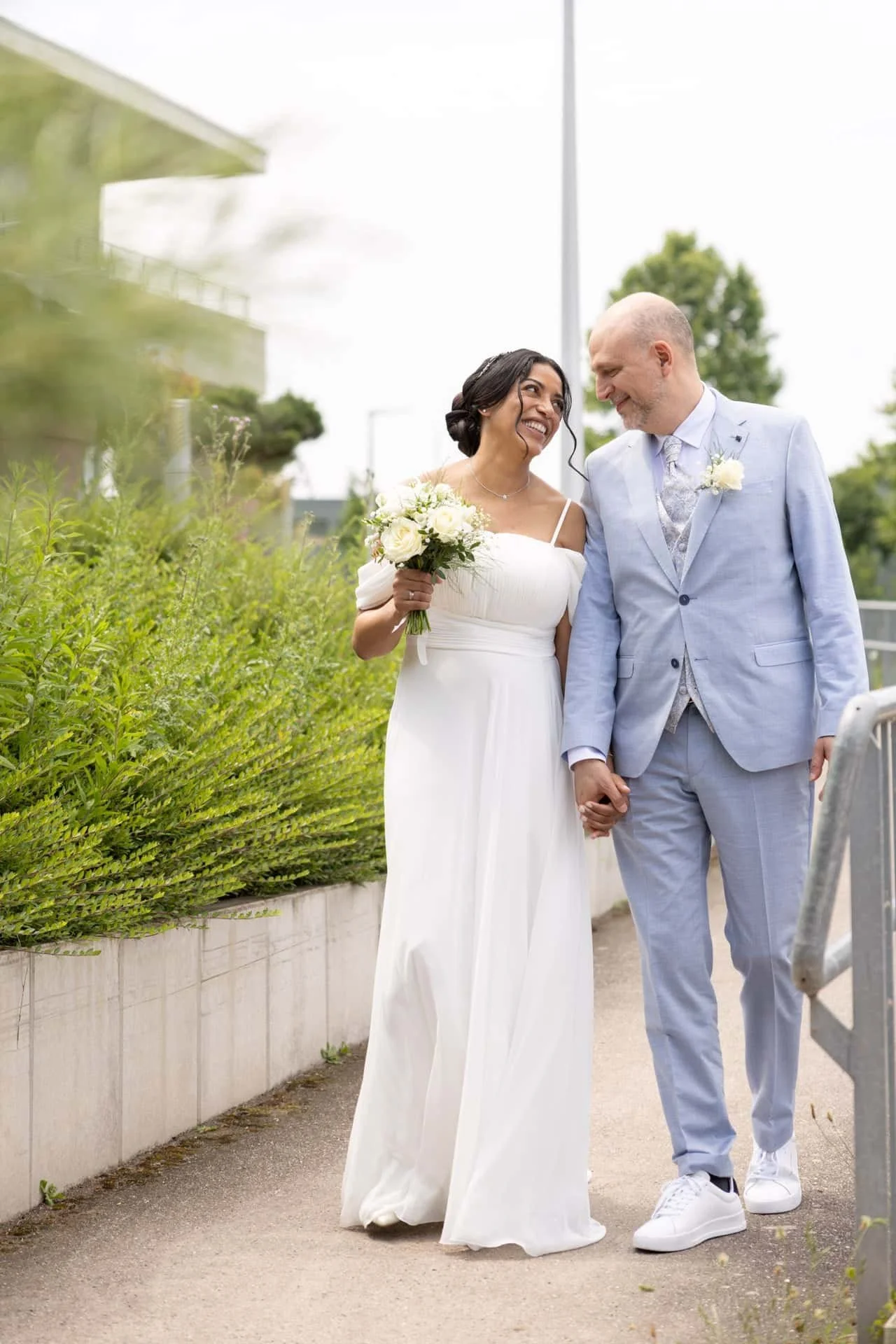 Un couple de mariés marchant main dans la main, la femme portant une robe blanche et tenant un bouquet, souriant vers l'homme qui porte un costume bleu clair.