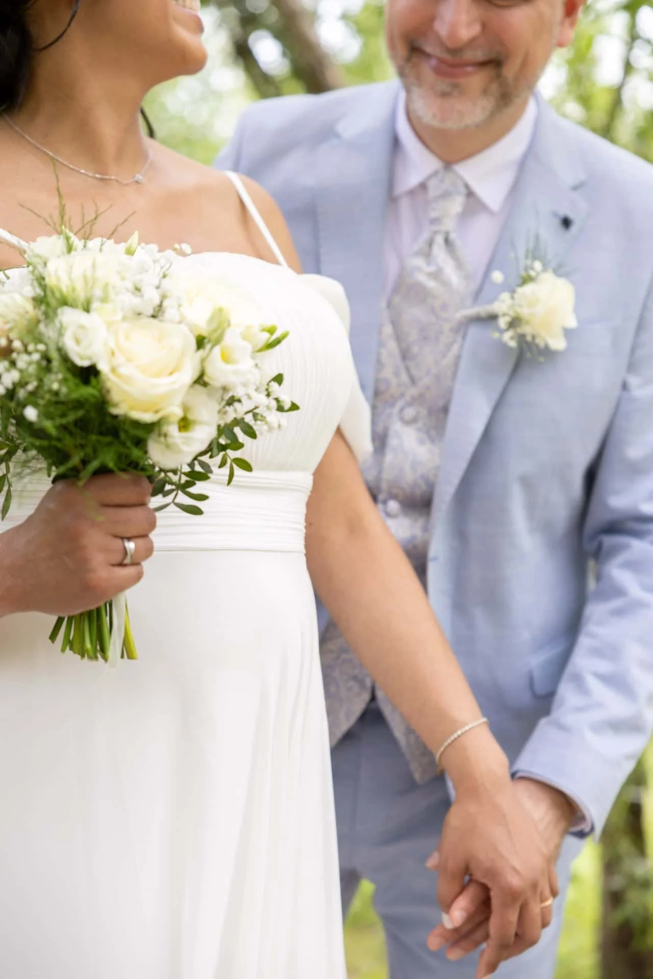 Un couple de mariés tenant la main lors d'un mariage en plein air, avec la femme en robe blanche tenant un bouquet de fleurs blanches, et le marié en costume bleu clair, tous deux souriants.