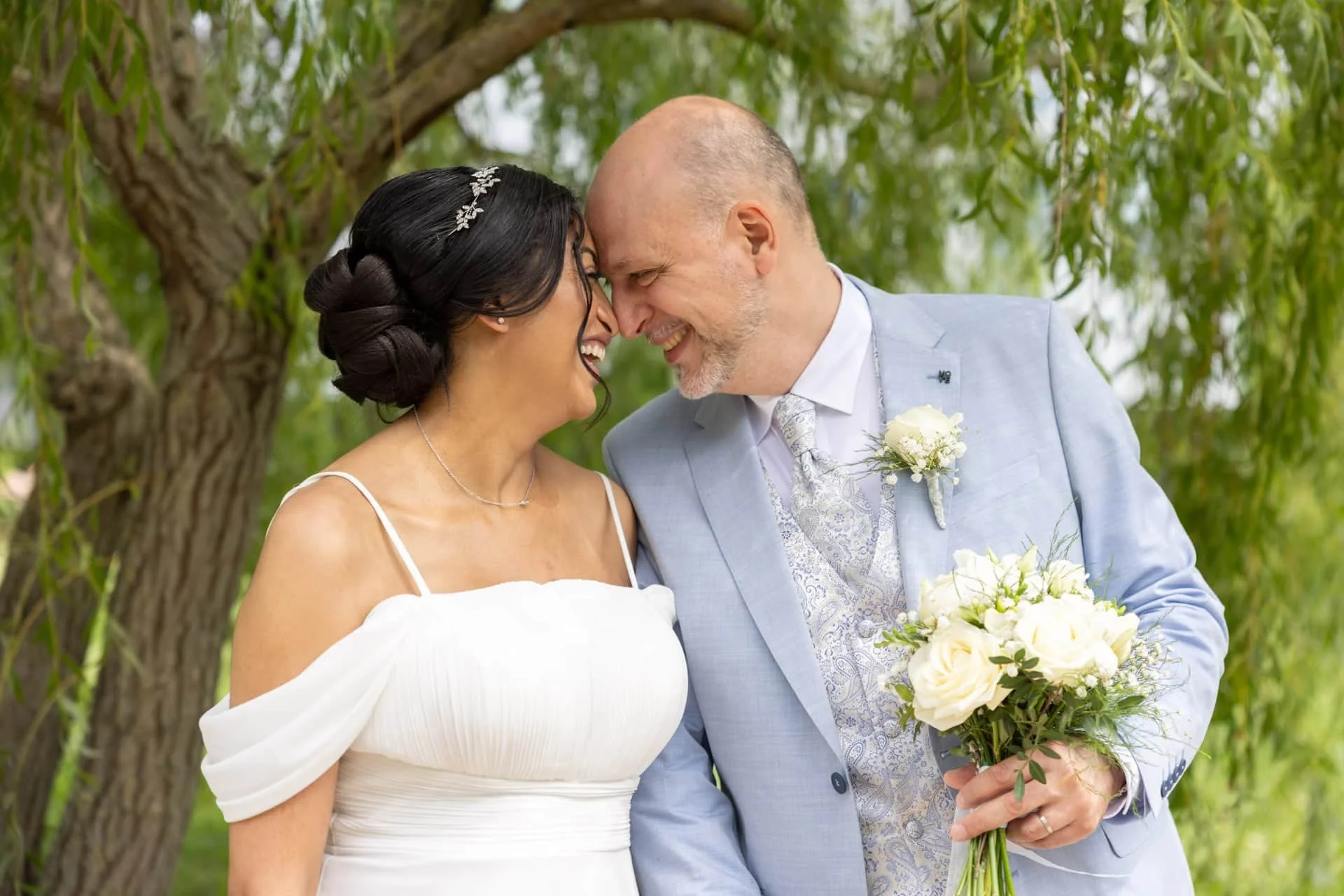 Un couple de mariage souriant, face à face, sous un arbre vert, la femme en robe blanche et le homme en costume clair, tenant un bouquet de fleurs blanches.