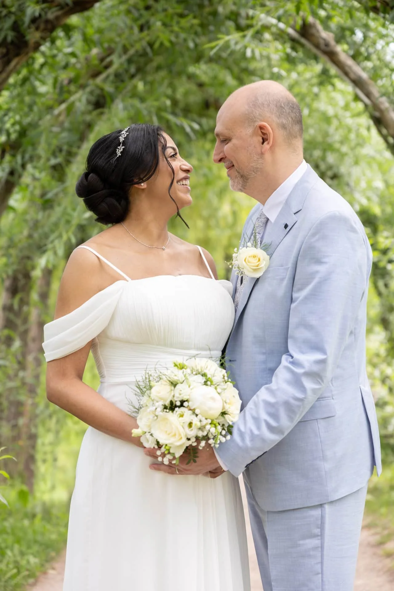 Un couple de mariés se regarde dans un cadre naturel, la femme en robe blanche et le homme en costume gris clair, tenant un bouquet de fleurs blanches.