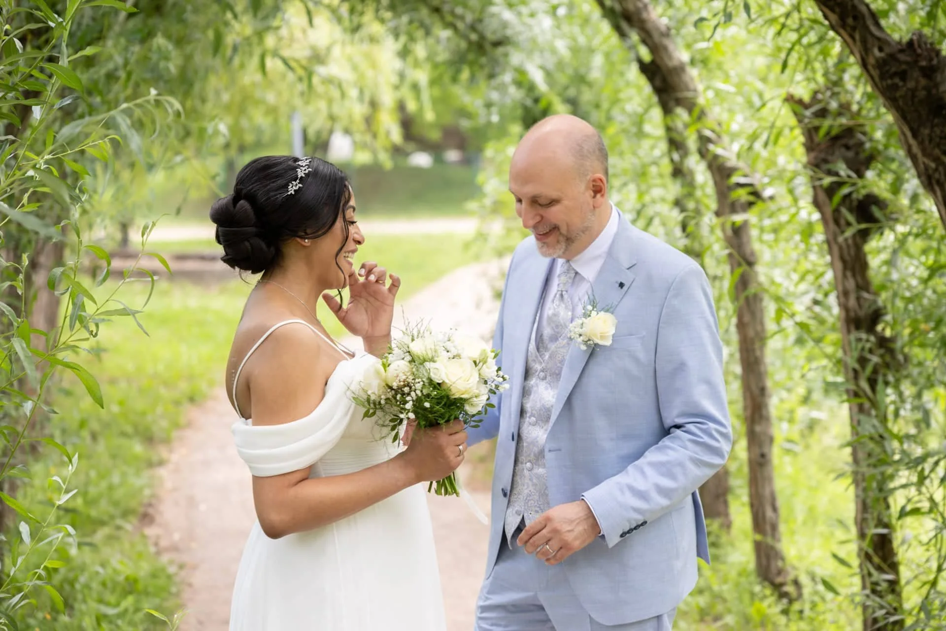 Un couple lors d'un mariage, homme et femme souriant, dans un cadre naturel avec des arbres, elle tenant un bouquet blanc.