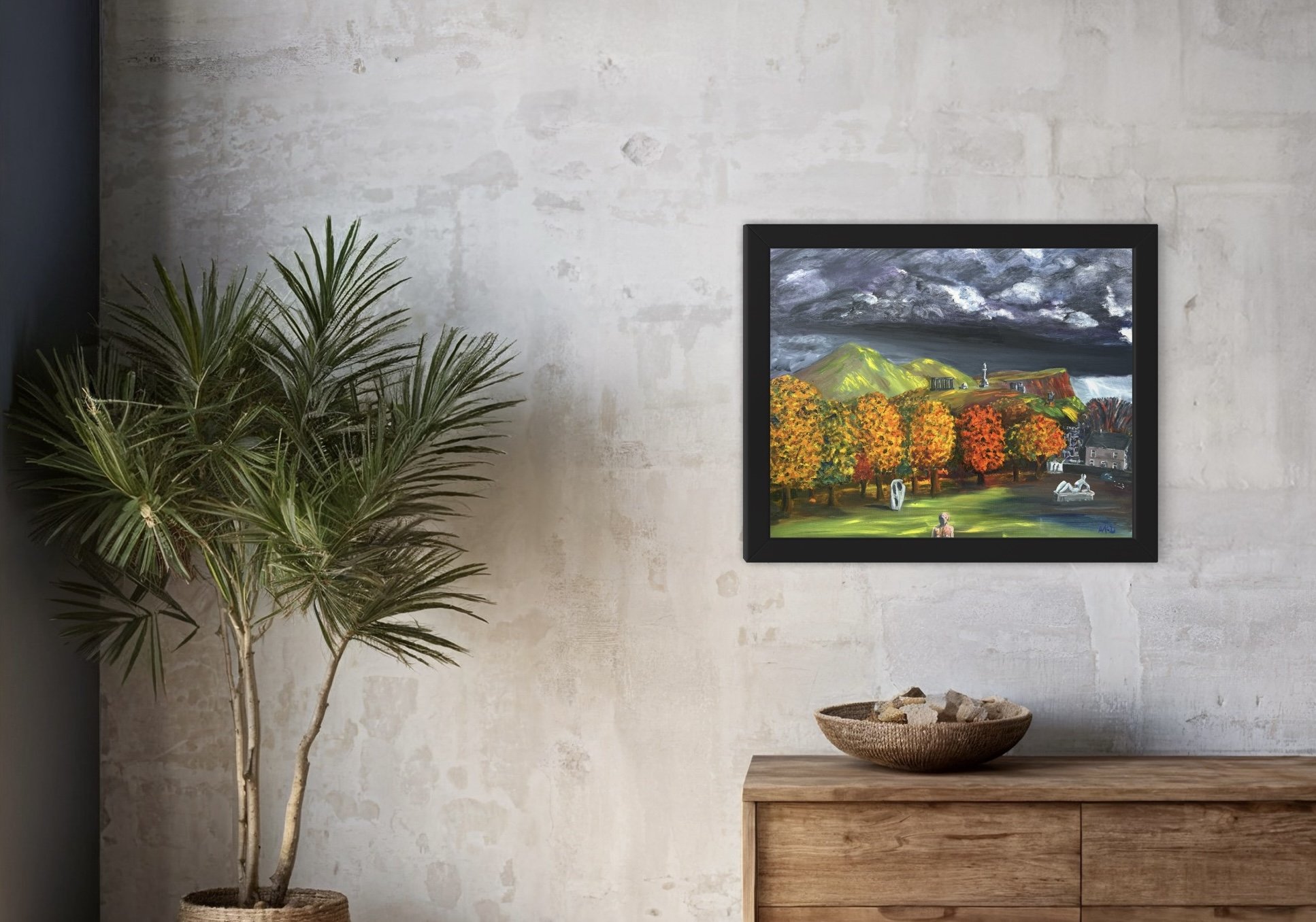 A room interior with a potted plant on the left, a wooden sideboard with a basket of rocks in the center, and a framed landscape painting on the wall showing mountains, trees with colorful fall foliage, and cloudy skies.