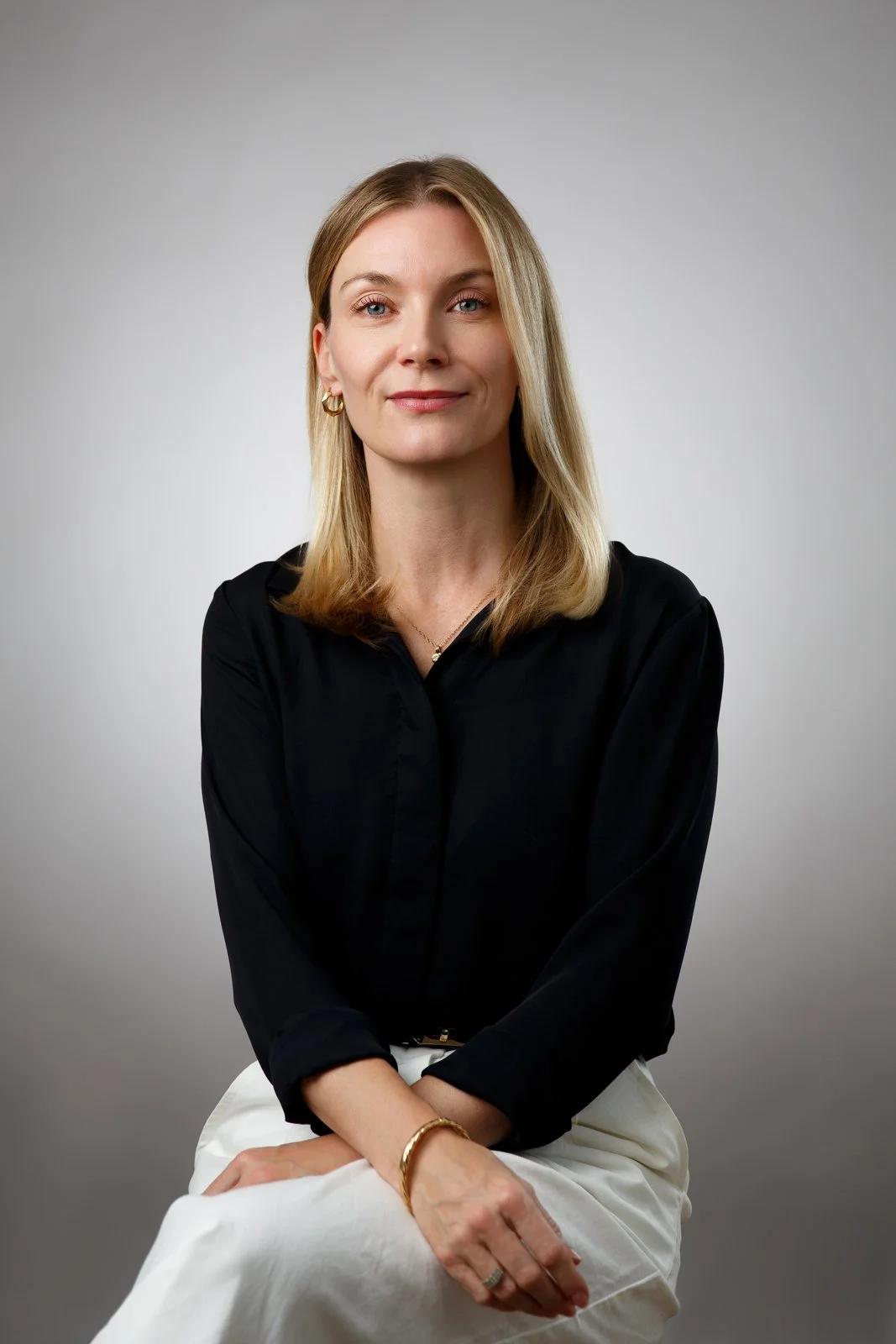Portrait of a woman with blonde hair wearing a black blouse and white skirt, sitting against a gray background.