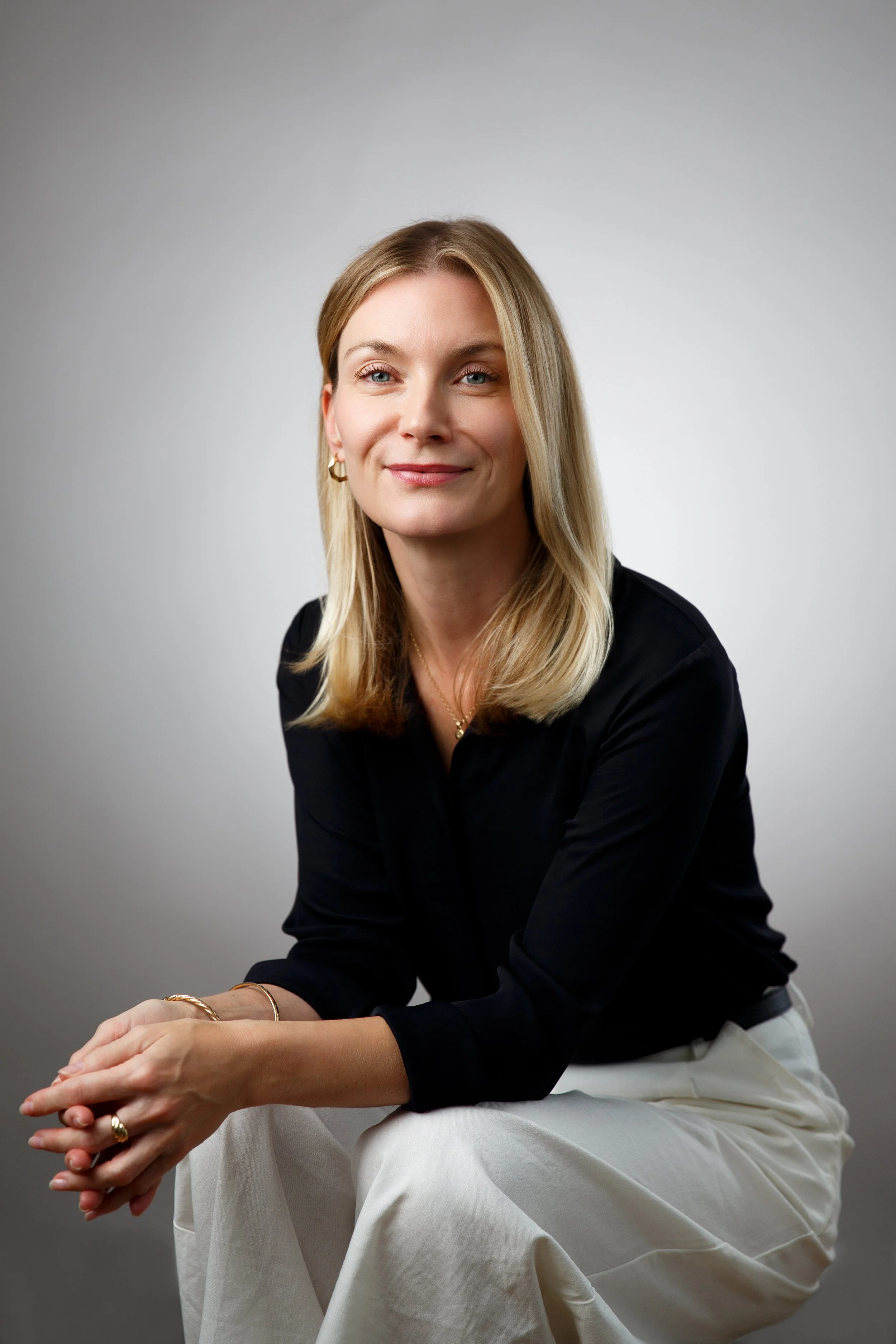 A woman with blonde hair wearing a black top and white pants, sitting against a plain background, smiling at the camera.