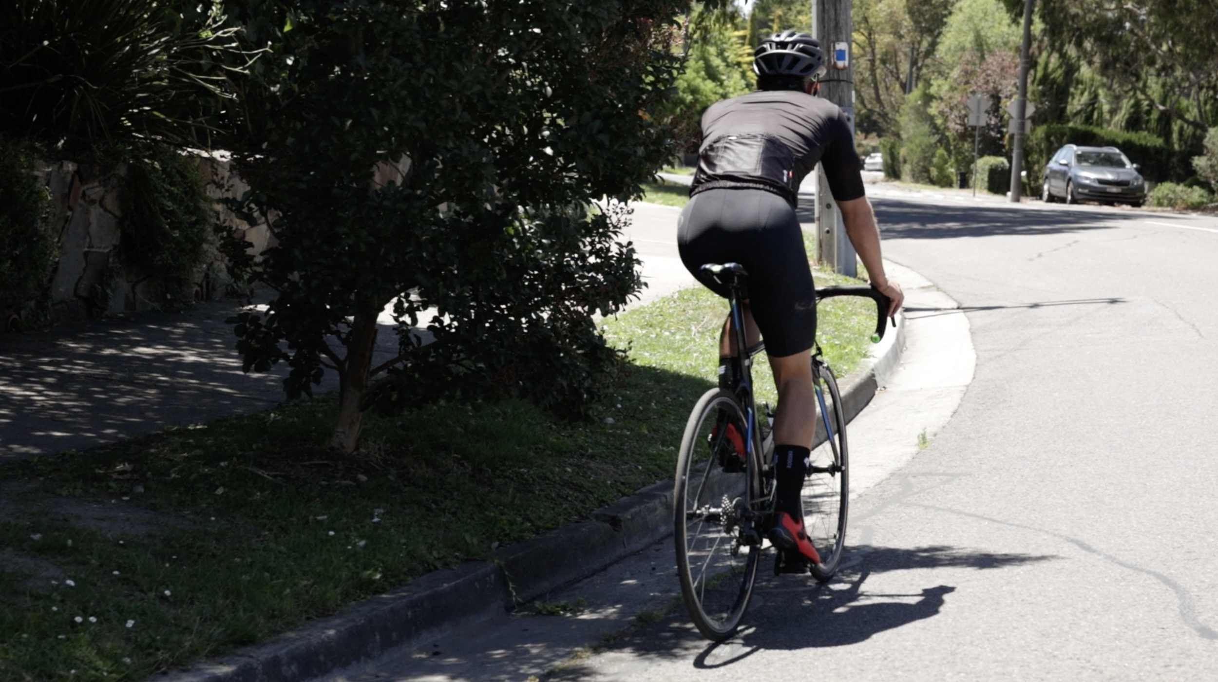 A person riding a road bicycle on a sidewalk near a tree and a street, wearing a helmet and cycling gear.