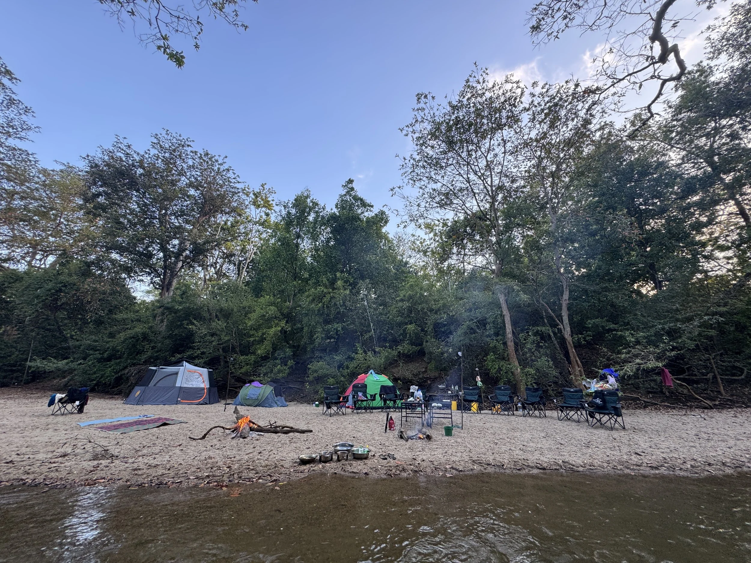 3 tents by the river with 10 camp chairs arranged neatly. A fire burns on the side and a folding table is seen in the middle.