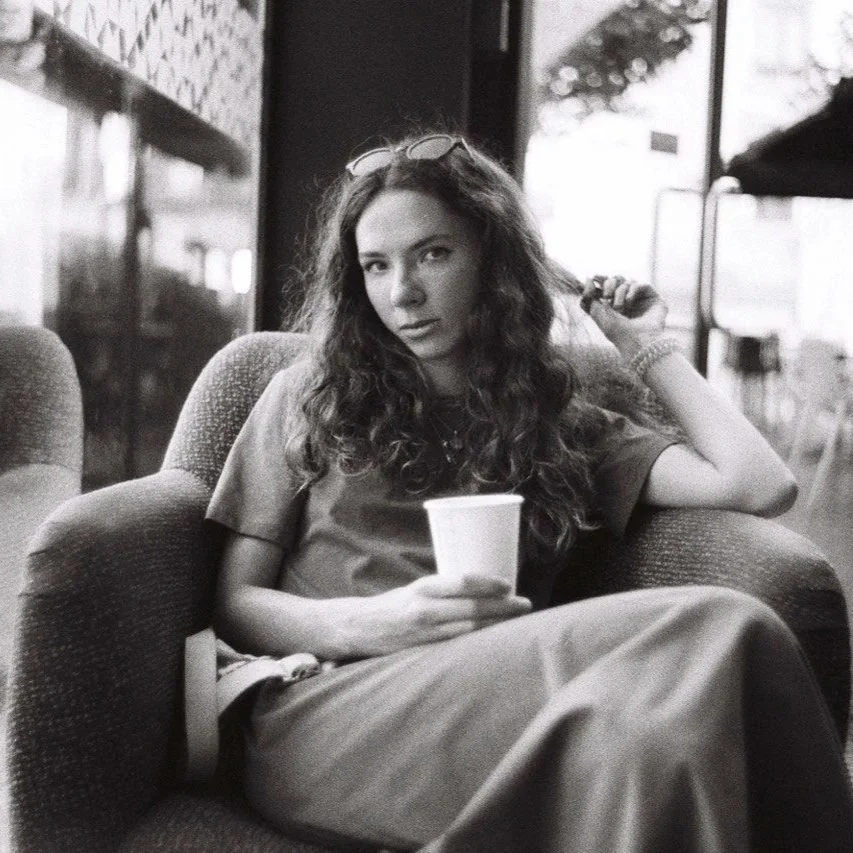 A young woman with long, curly hair sitting on a cushioned armchair in a coffee shop, holding a cup, with sunglasses on her head and casual clothing.