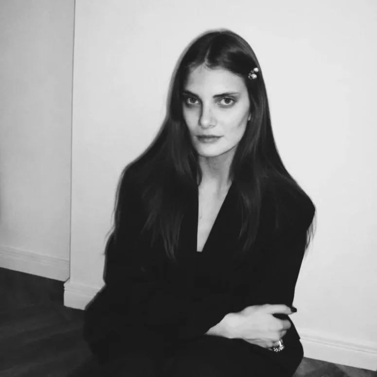 Black and white photo of a woman with long dark hair, sitting on the floor, wearing a dark top, with a neutral facial expression, against a plain white wall.