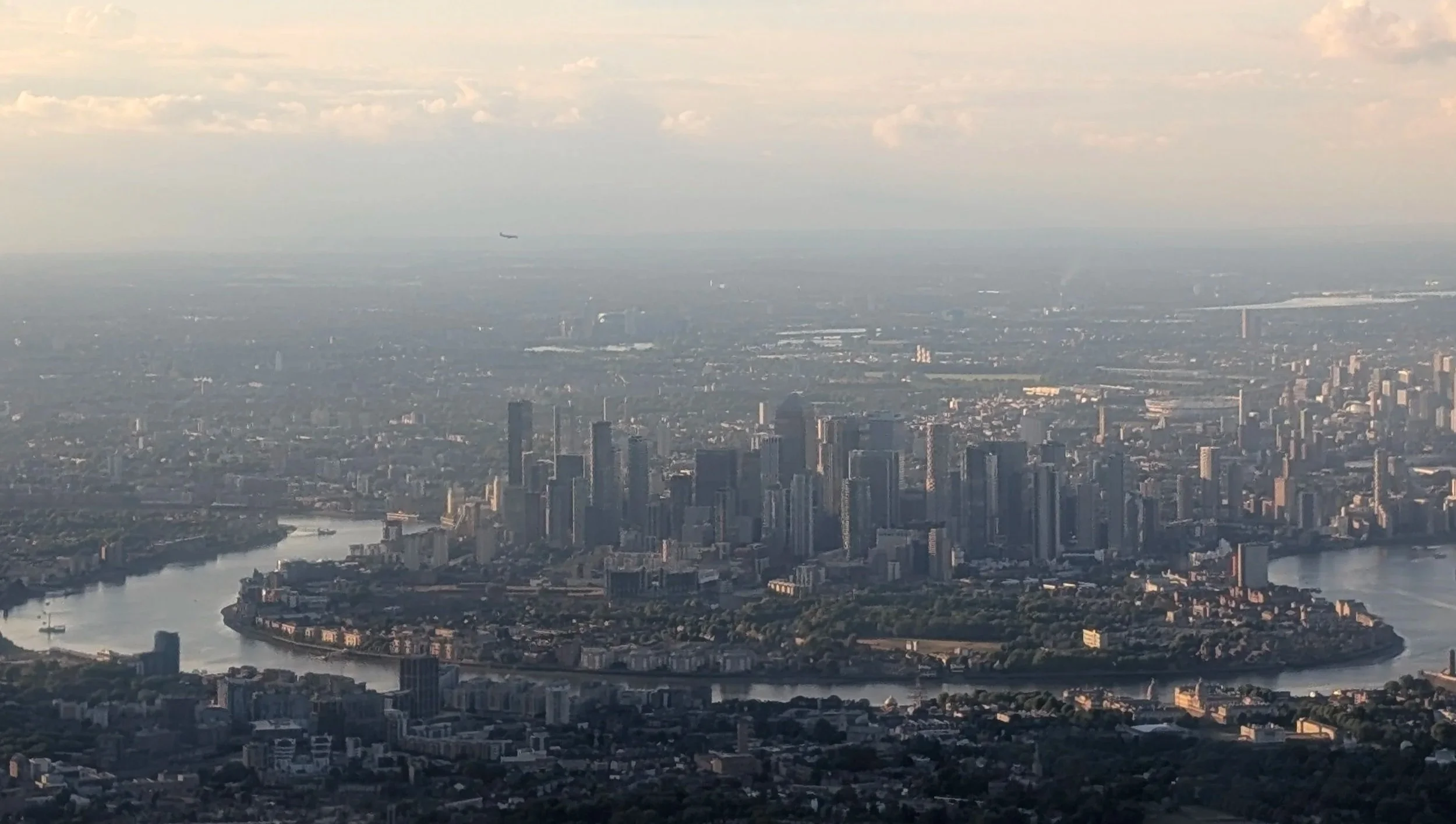 Aerial view of London skyline featuring numerous tall skyscrapers, river winding through the city, and a distant airplane in the sky.