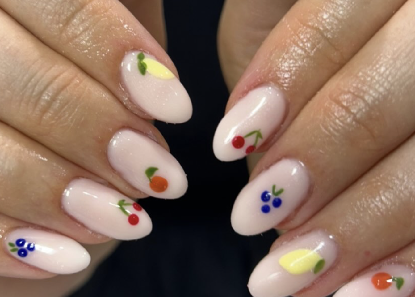 Close-up of manicured fingernails painted in a light pink color, decorated with small, colorful fruit designs including cherries, blueberries, raspberries, a peach, and lemon slices.