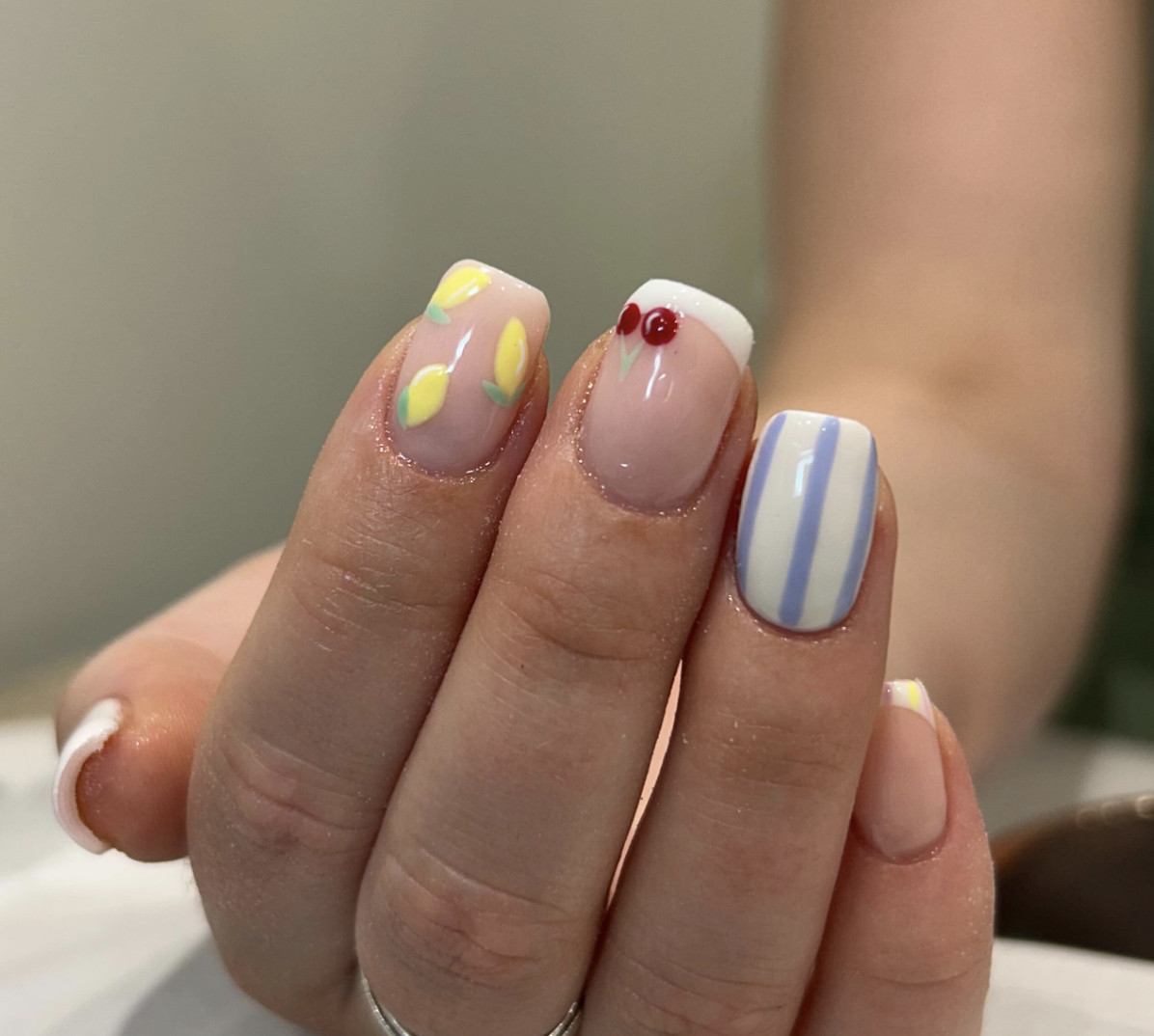 Close-up of a hand with manicured nails featuring colorful, spring-themed designs including lemons, cherries, and pastel stripes.