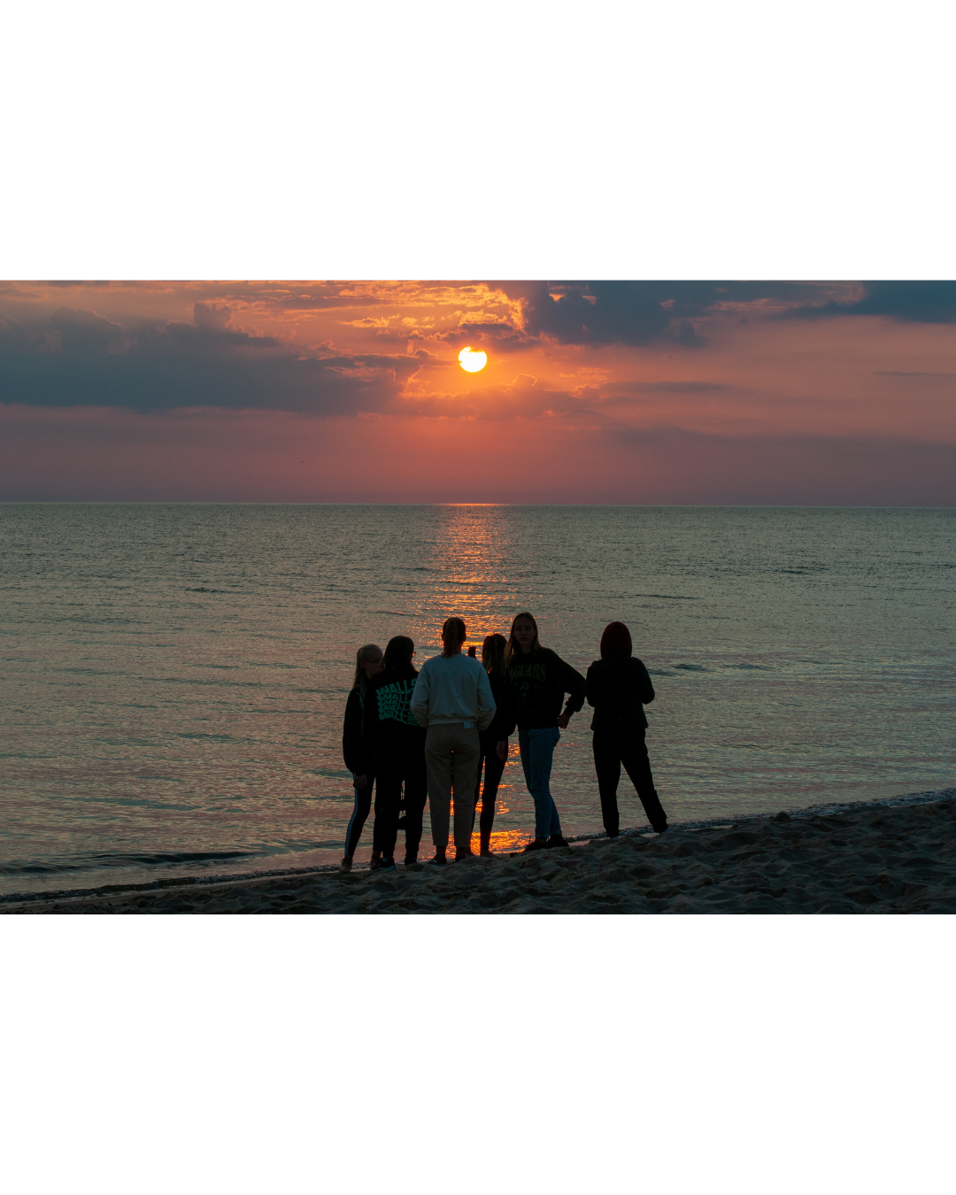 Group of people standing on a beach at sunset, with the sun partially covered by clouds over the ocean.