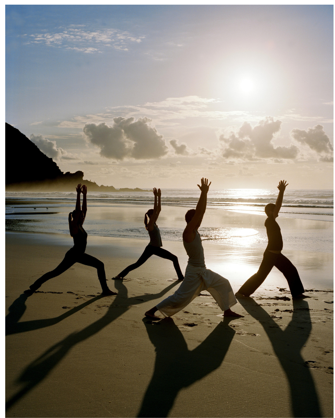 A group of four people practicing yoga on a sandy beach at sunset, performing warrior poses with shadows cast on the sand, with the ocean and cloudy sky in the background.