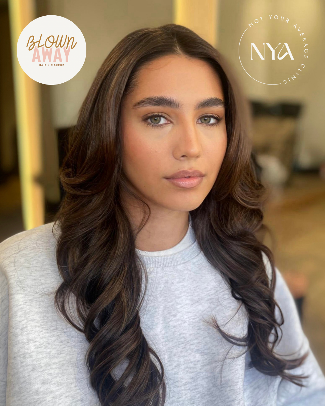 Woman with long, wavy brown hair wearing a gray sweatshirt, with logos for "Blown Away Hair + Makeup" and "Not Your Average Clinic (NYA)" displayed.
