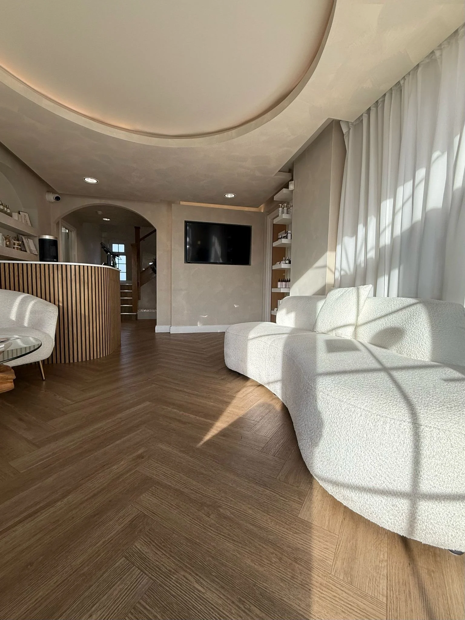 Modern interior with light wood flooring, a curved white sofa, a wooden reception desk, a flat-screen TV on the wall, shelves with products, and large windows with white curtains.