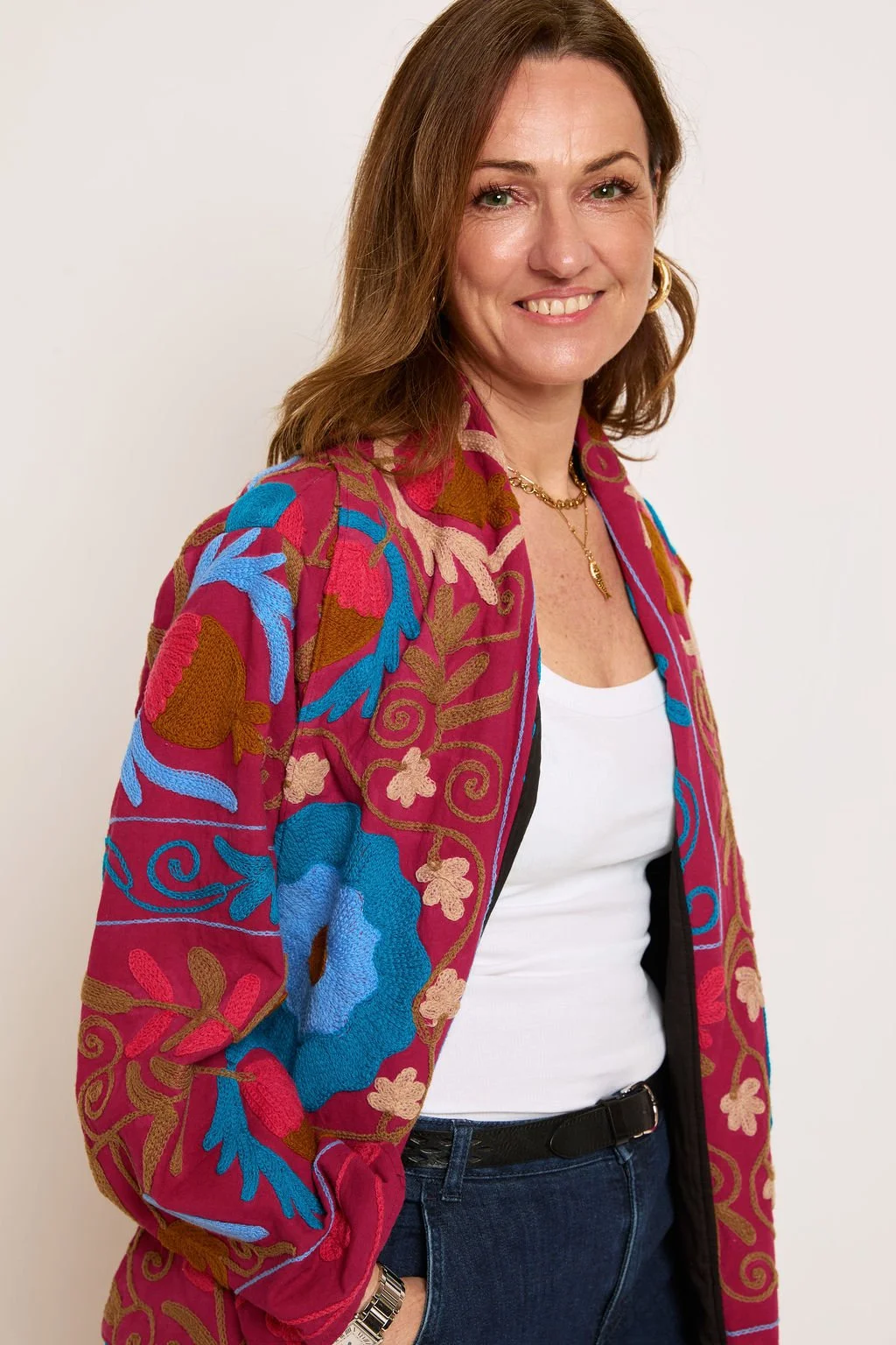A woman with shoulder-length brown hair, wearing a colorful embroidered jacket over a white top, black belt, blue jeans, and gold jewelry, smiling at the camera against a plain white background.