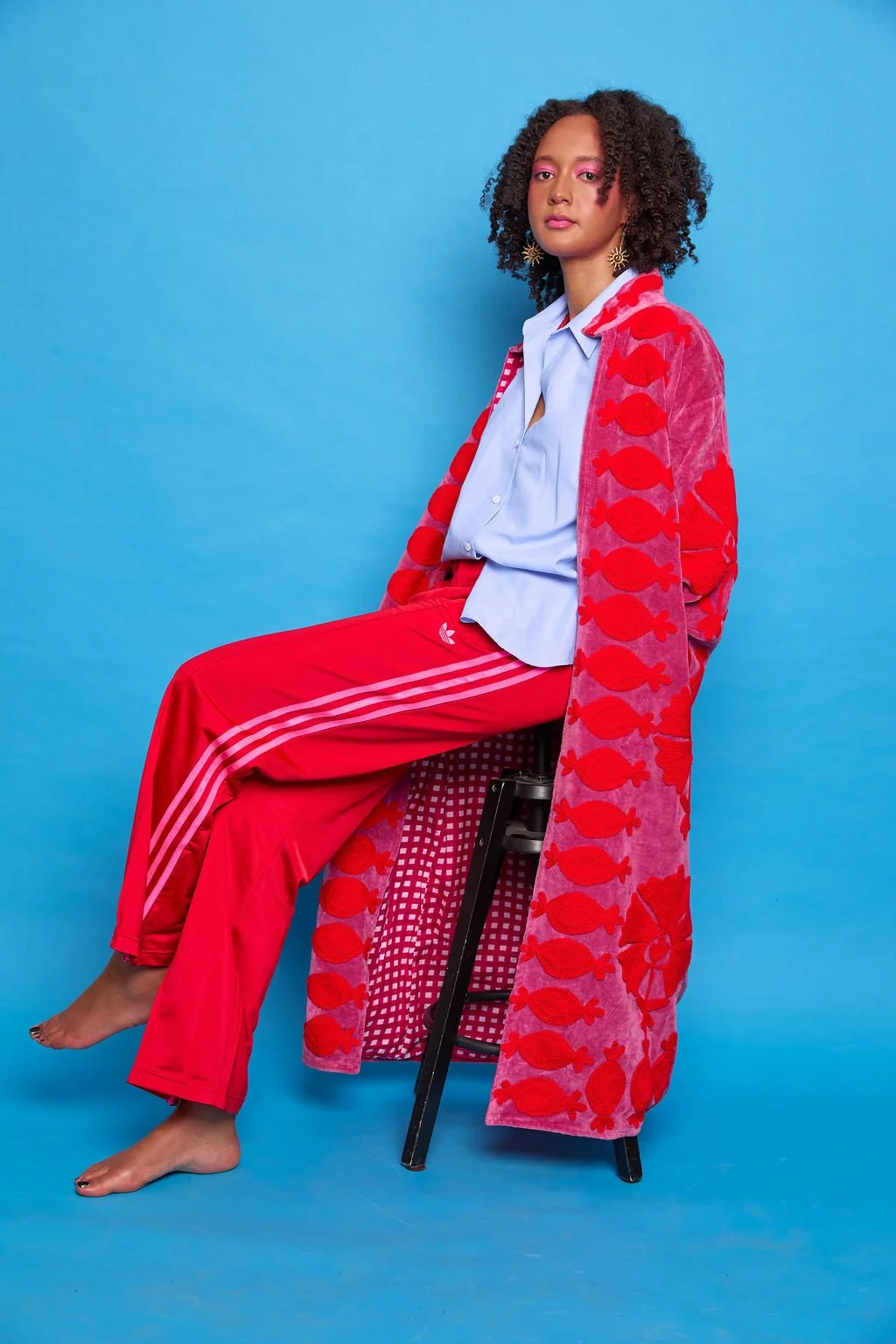A woman sitting on a stool against a blue background, wearing a colorful outfit with a red fish-patterned shrug, a light blue shirt, and red Adidas track pants.