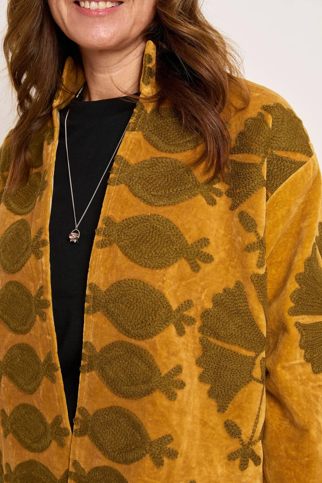 Close-up of a woman wearing a mustard yellow jacket with green embroidery of bird shapes, black shirt, and a silver necklace with a pendant.