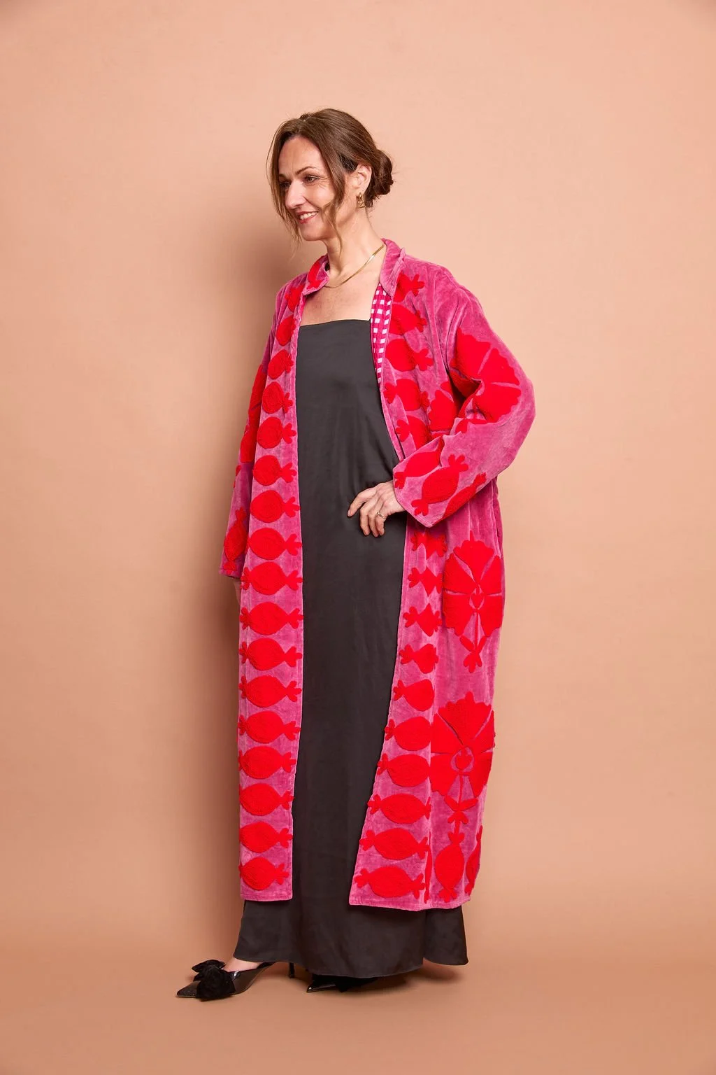 Woman in a long, black dress and a pink robe with red fish patterns, standing against a beige background.
