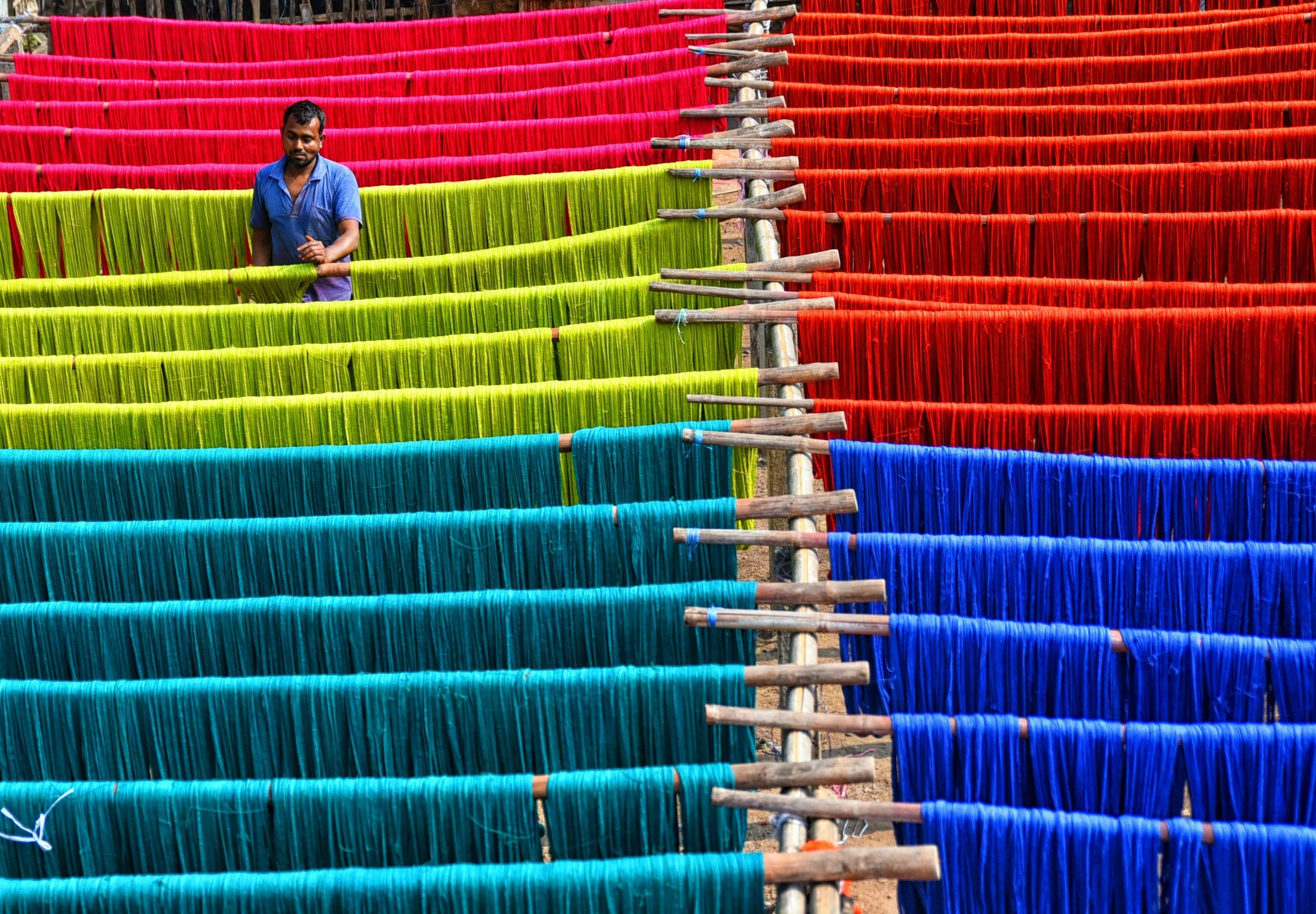 A man in a blue shirt stands among colorful rows of dyed yarns hanging on wooden rods in an outdoor area, with colors ranging from green, yellow, red, to blue.
