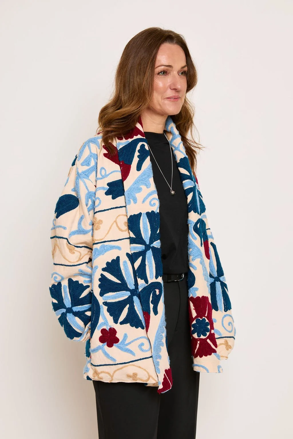 Woman with brown hair wearing a colorful embroidered jacket, black top, and black pants against a plain white background.