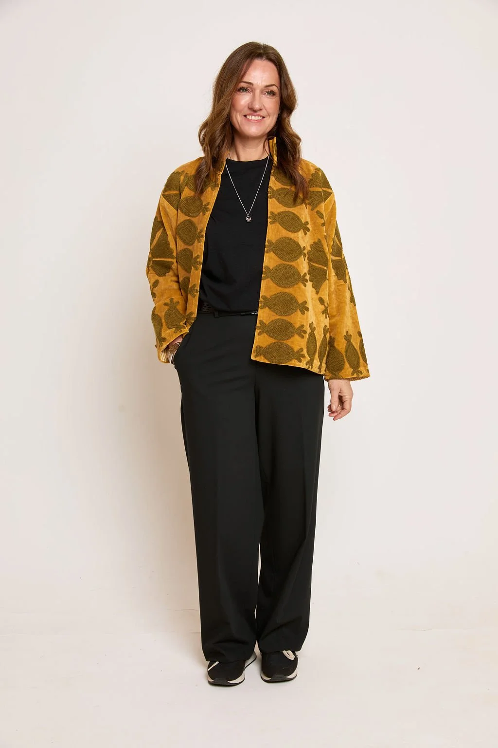 A woman with shoulder-length brown hair smiling, wearing a yellow-brown jacket with fish and rabbit patterns, black top, black pants, and sneakers, standing against a plain white background.