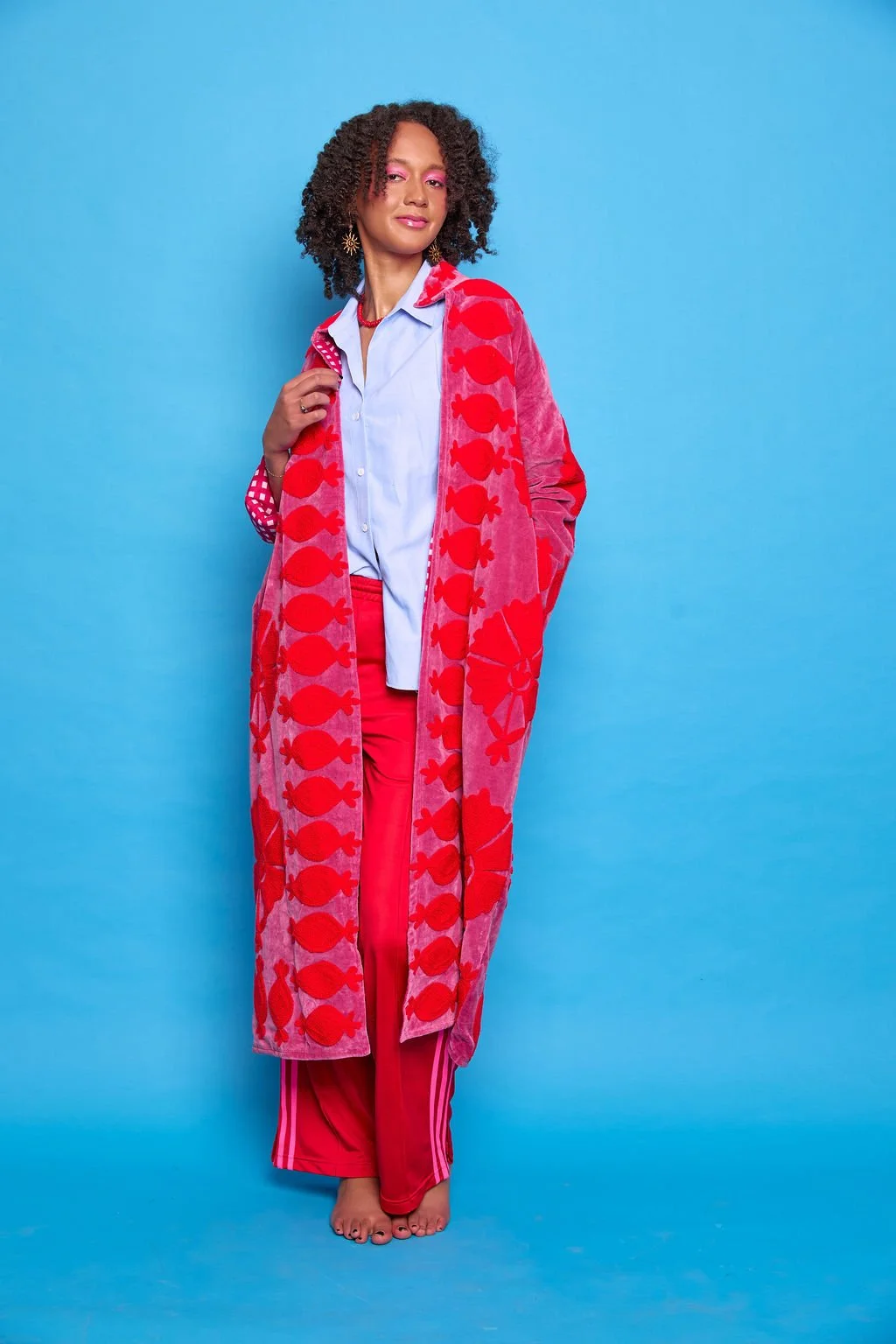 Woman standing barefoot against a blue background, wearing a blue shirt, red pants, and a pink and red patterned coat.