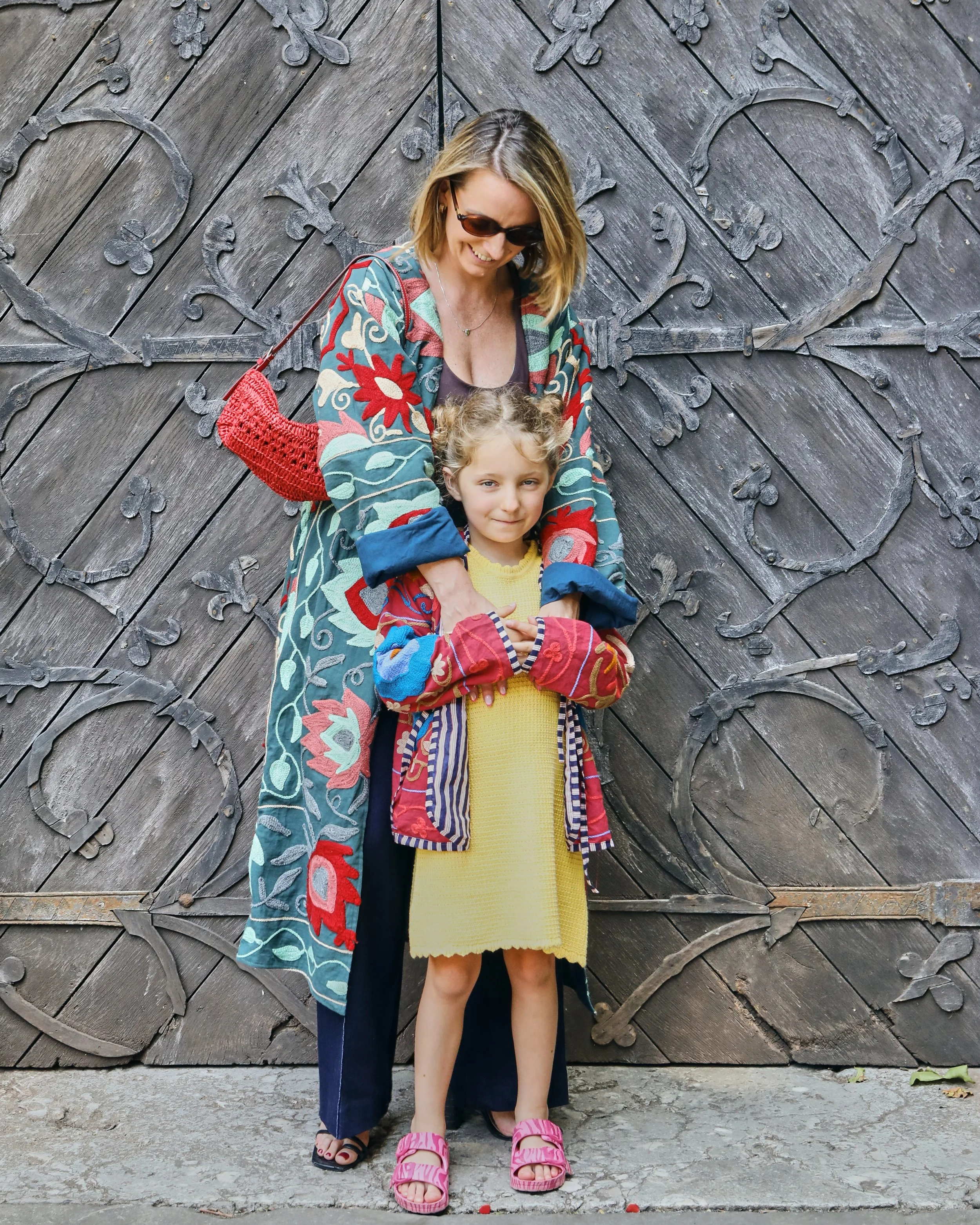A woman and a young girl standing in front of a decorative wooden door. The woman is wearing sunglasses, a colorful quilted coat, and flip-flops, with a red bag on her arm. The girl is wearing a yellow dress, a multicolored jacket, and pink sandals.