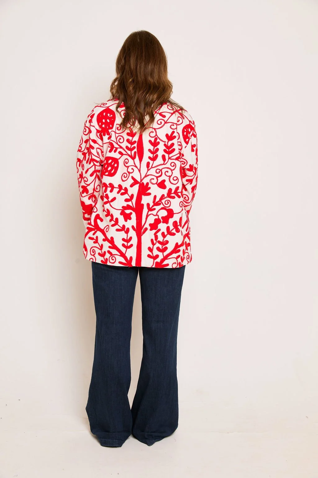 Back view of a woman with wavy brown hair wearing a white coat with red floral and vine patterns and dark wide-leg jeans, standing against a plain white background.