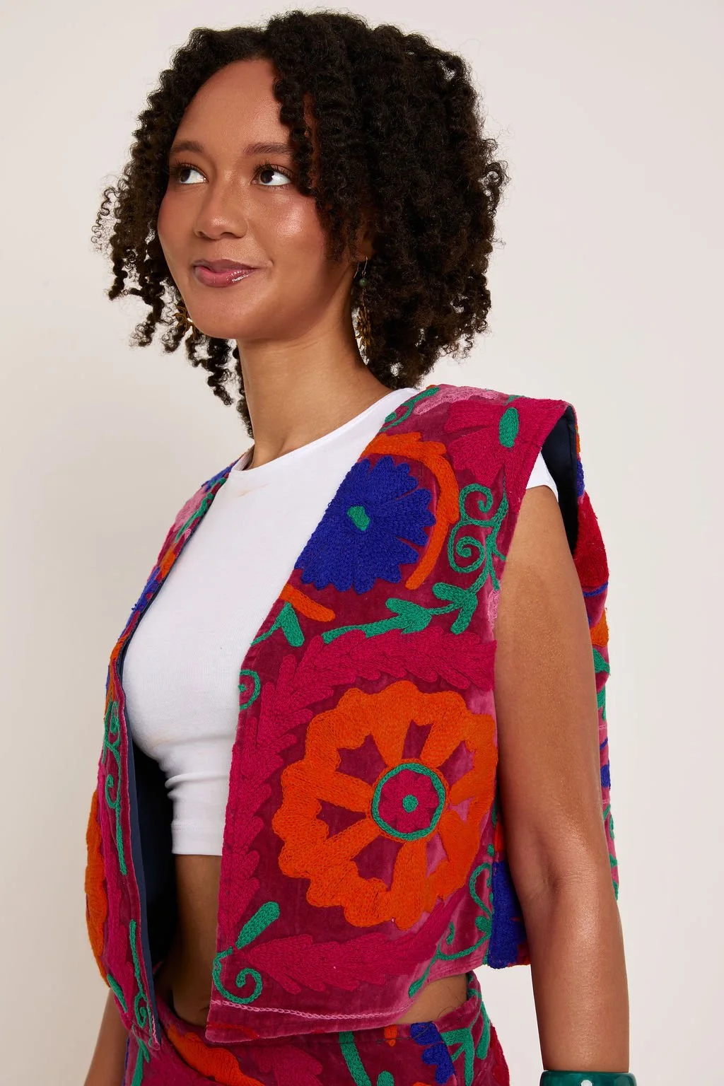 A woman with curly hair and earrings wearing a white top and a sleeveless, colorful embroidered vest with floral patterns.