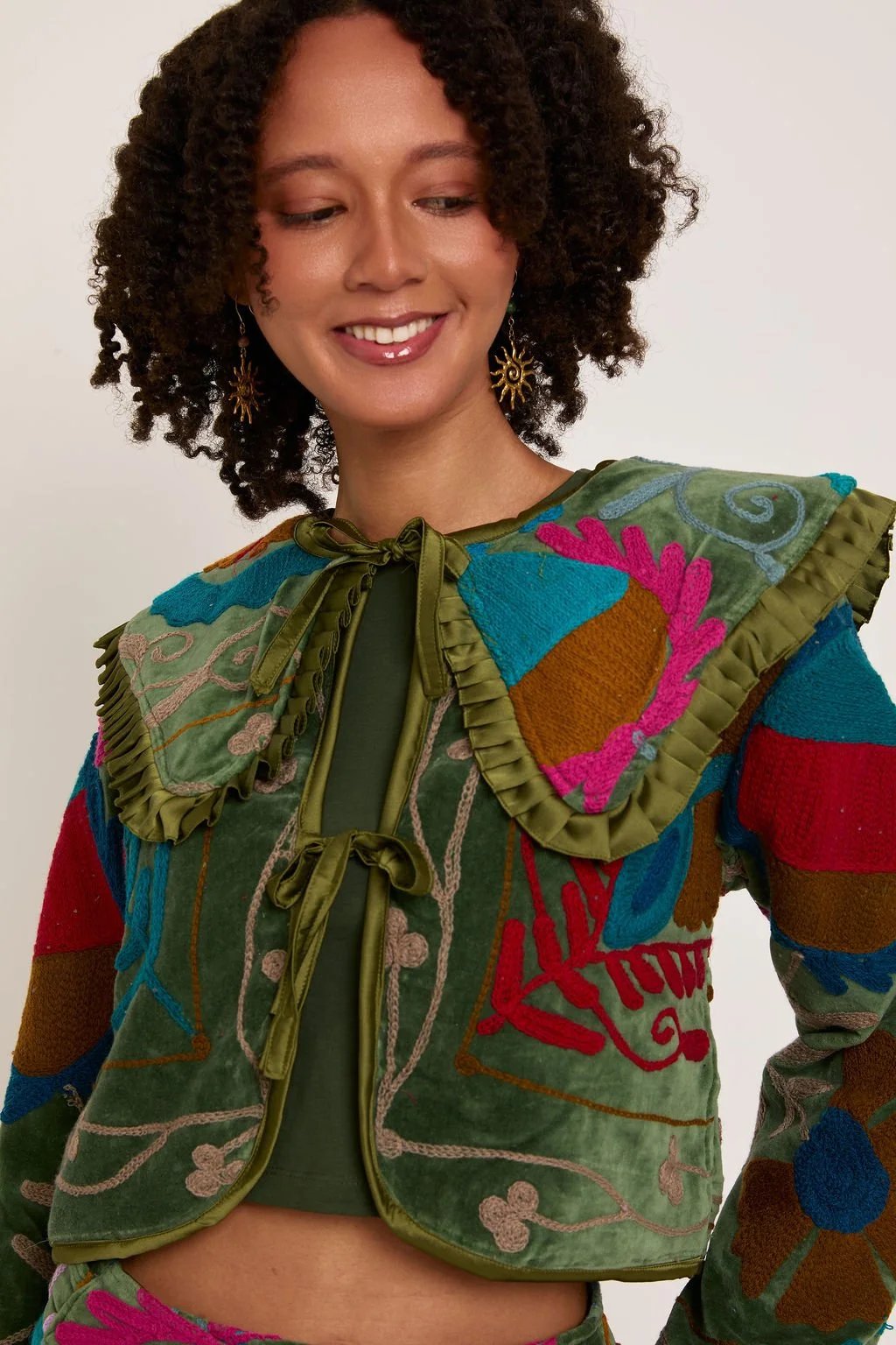 A woman with curly hair wearing a colorful embroidered jacket with large shoulder pads and green ribbon tie, smiling softly.