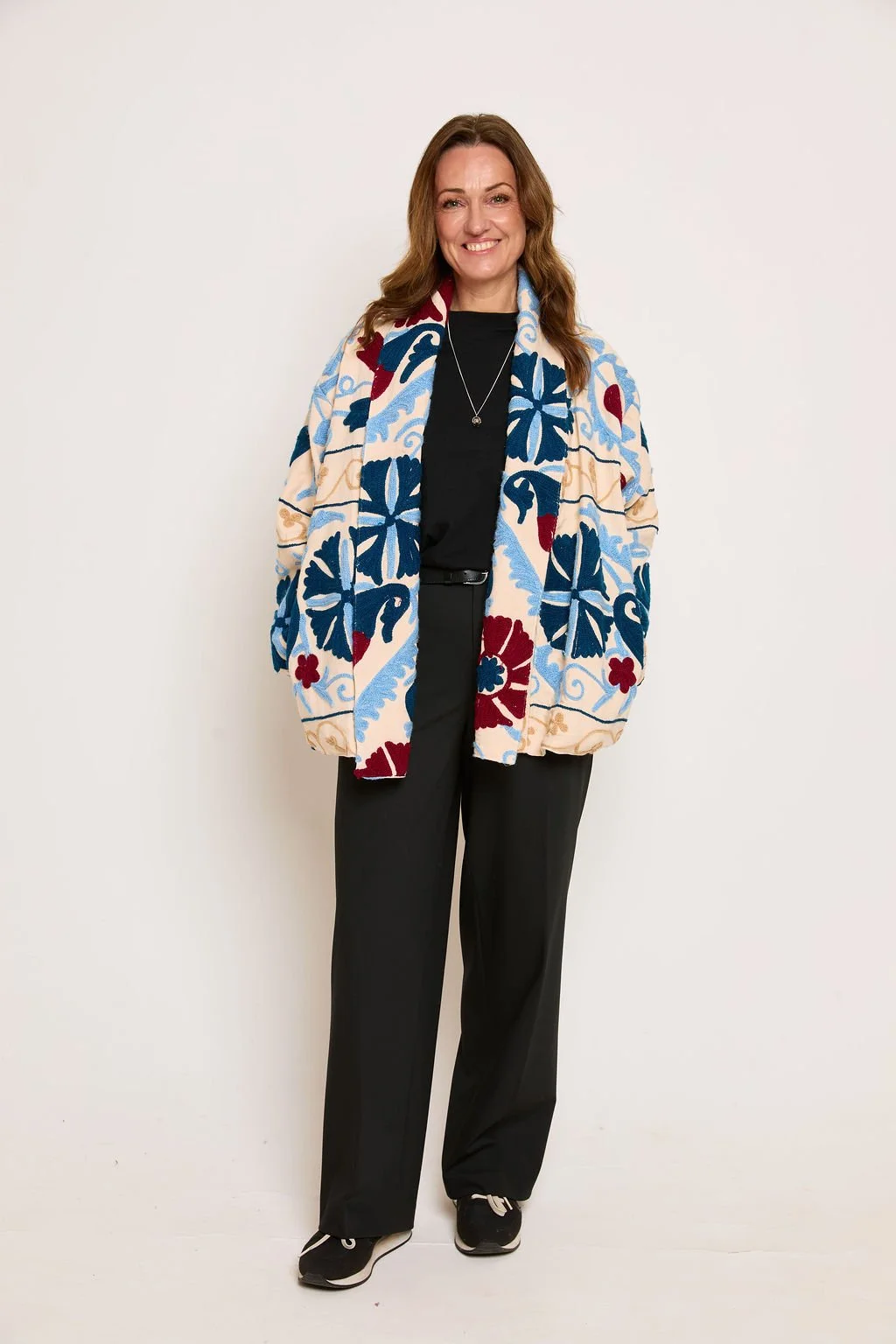 A woman with brown hair wearing a colorful floral-patterned jacket, black pants, and black sneakers, standing against a plain white background, smiling.