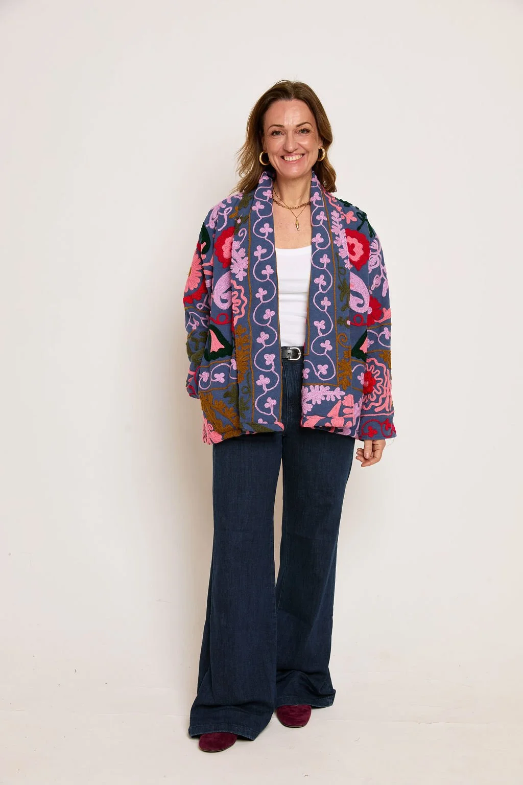 A woman smiling, wearing a colorful floral embroidered jacket, white top, dark wide-leg jeans, and red shoes, standing against a plain light background.