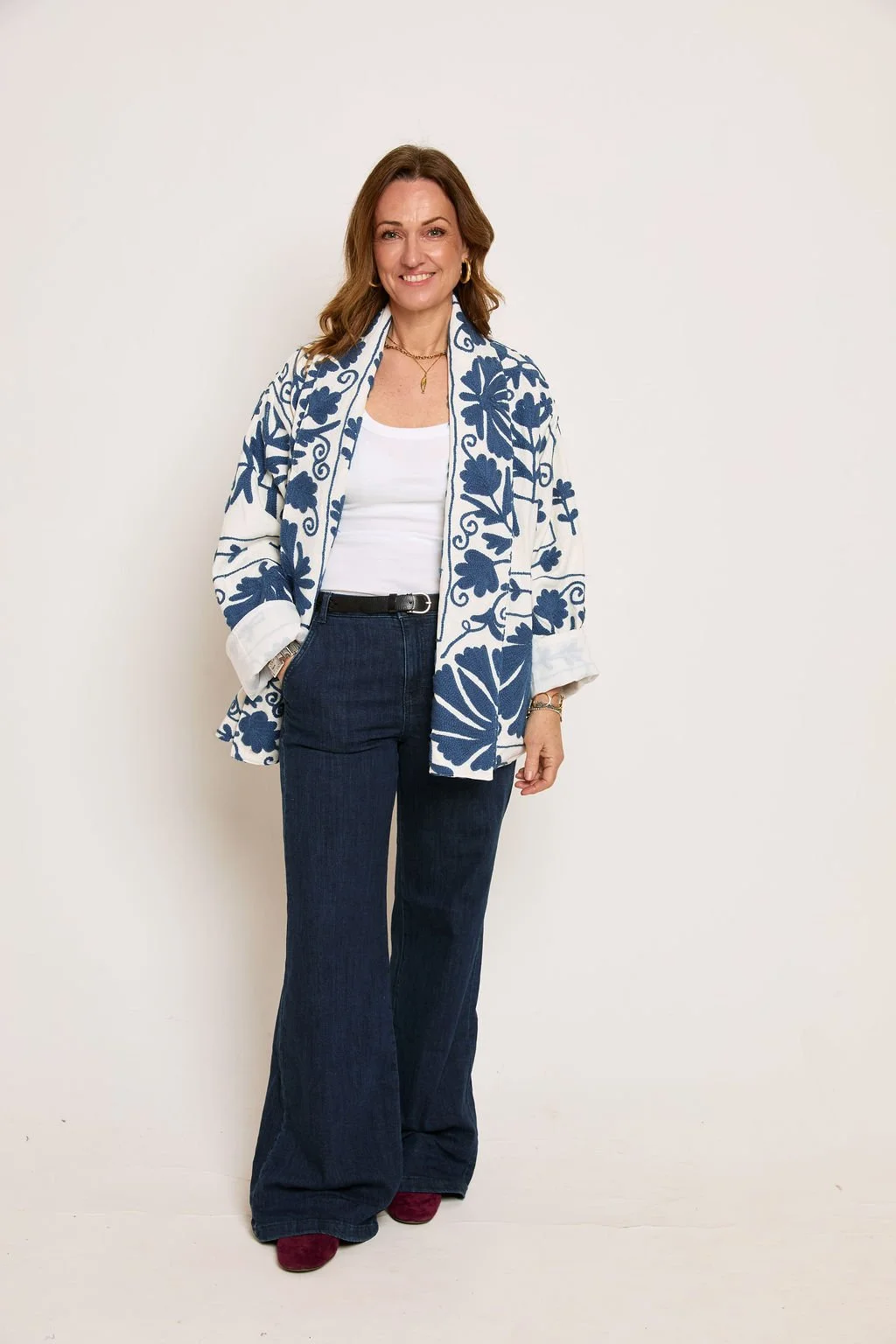 Woman with brown hair smiling, wearing a white top, dark wide-leg jeans, a white and blue floral jacket, pink shoes, and gold jewelry, standing against a plain white wall.
