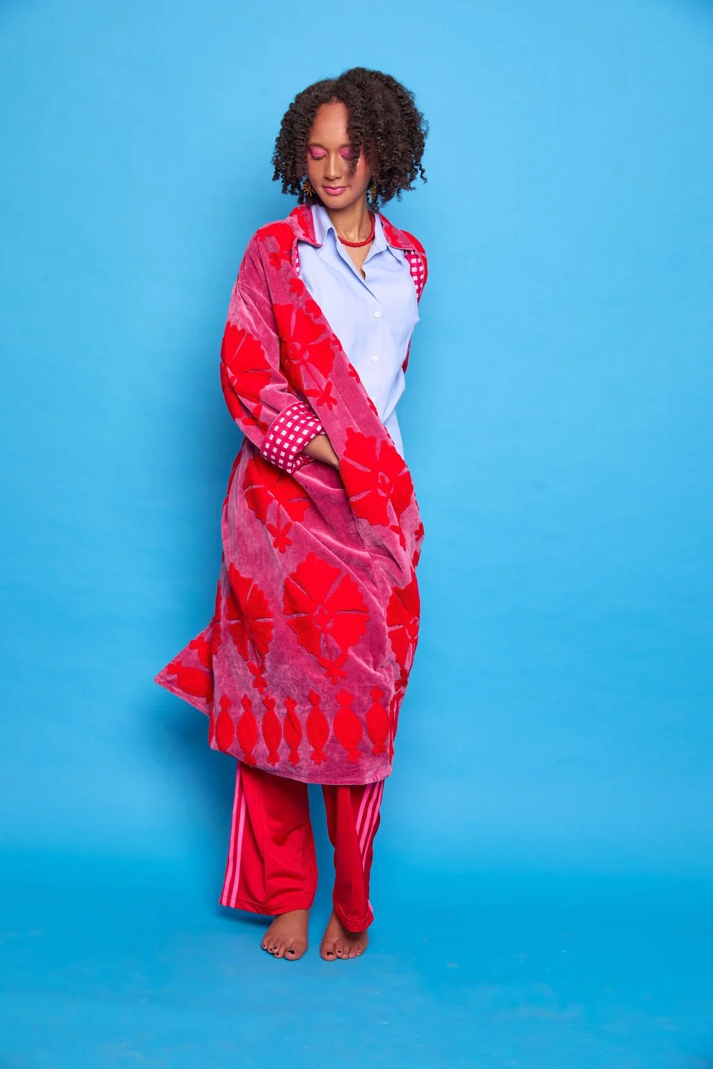 A woman with curly hair standing barefoot against a blue background, wearing pink eye makeup, a blue shirt, and a colorful red and pink patterned oversized coat.