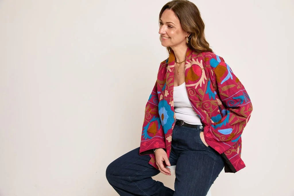 A woman with brown hair in a colorful, embroidered jacket, white top, and dark jeans, sitting against a plain white background, smiling.