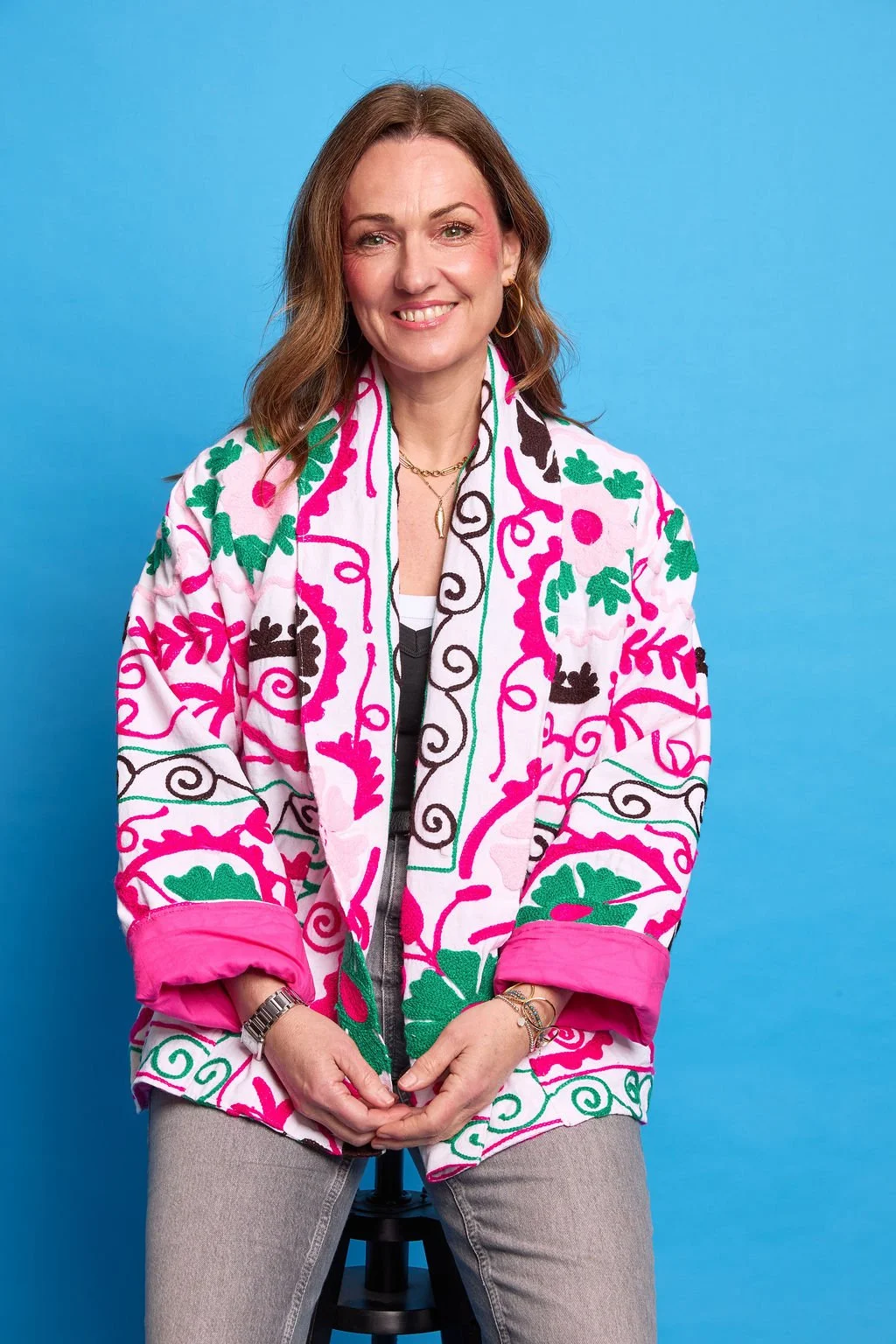 A woman with shoulder-length brown hair, wearing gold hoop earrings, a gold necklace, and a colorful embroidered jacket with pink, green, black, and white patterns, sits against a blue background smiling at the camera.