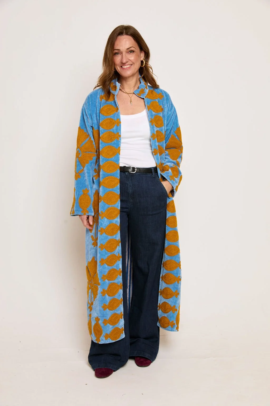 A woman with shoulder-length brown hair smiling, wearing gold hoop earrings, a gold necklace, a white top, dark blue wide-leg jeans, a black belt, and a long blue and orange patterned coat with fish designs, standing against a plain white wall.