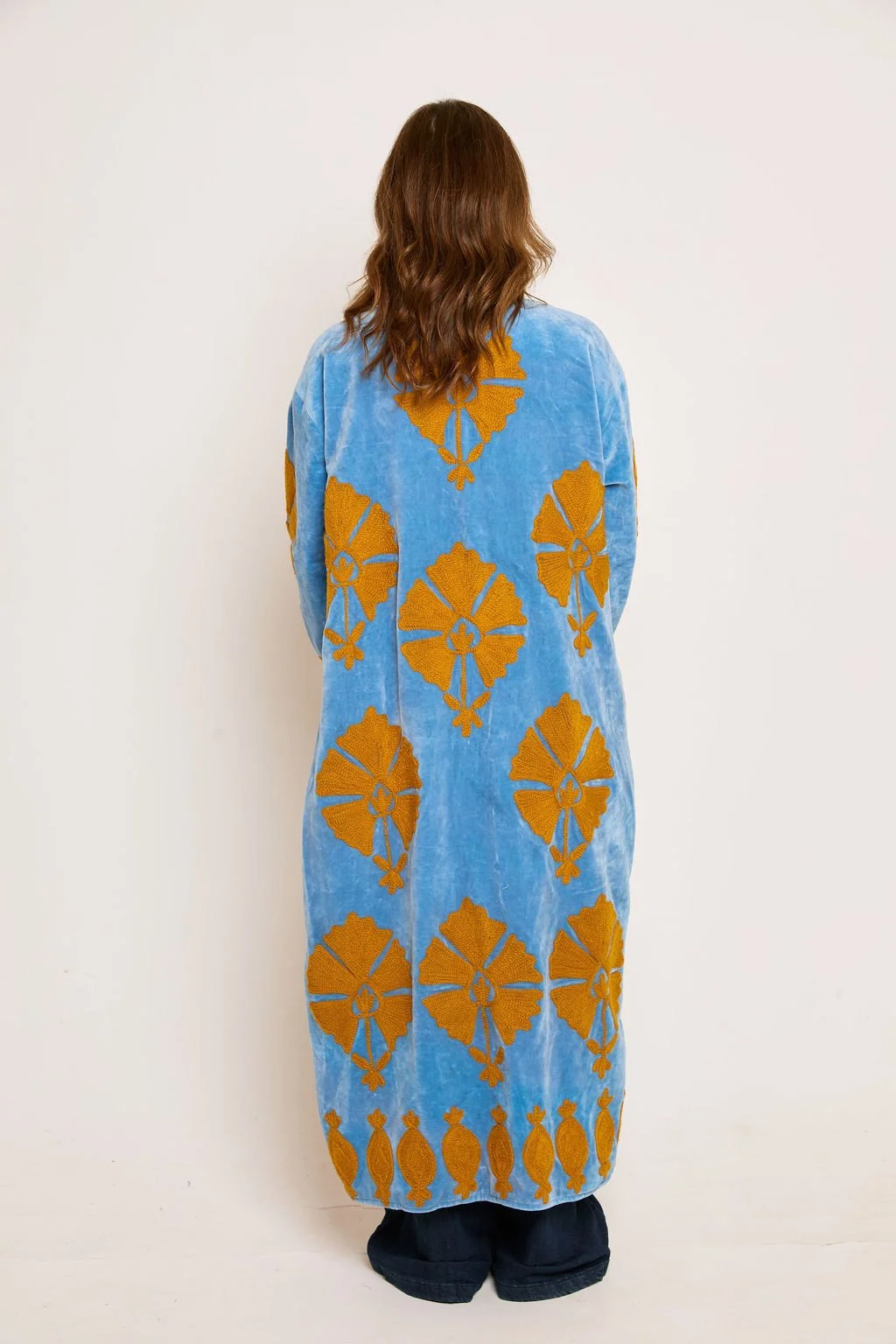 Back view of a woman wearing a long, light blue robe with large orange floral patterns and smaller orange designs at the bottom, standing against a plain white background.
