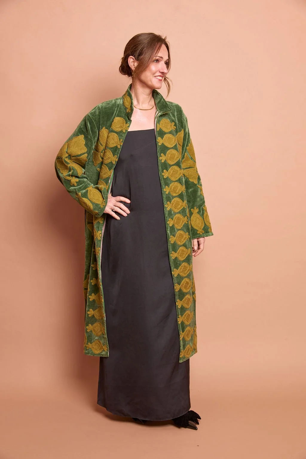A woman is wearing a black dress layered with a green velvet coat that has a repeating pattern of yellow fish. She is standing against a plain beige background, with her right hand on her hip and smiling.