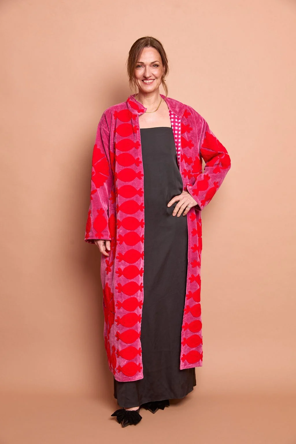 A woman standing against a beige background, wearing a pink bathrobe with red fish patterns, over a black dress, and black shoes.