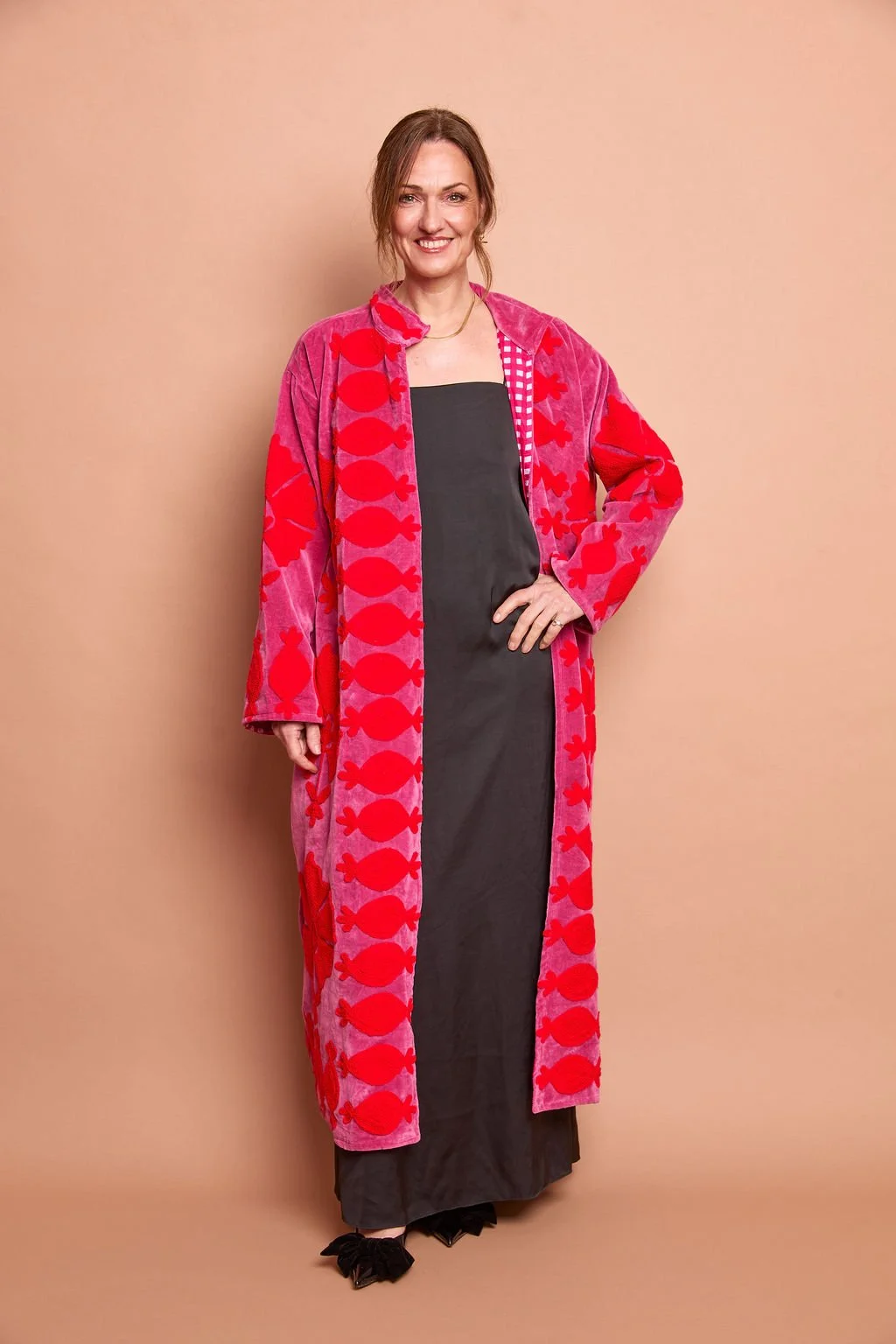 Woman standing against a beige background, wearing a long, pink and red patterned coat over a black dress, with her right hand on her hip and smiling.