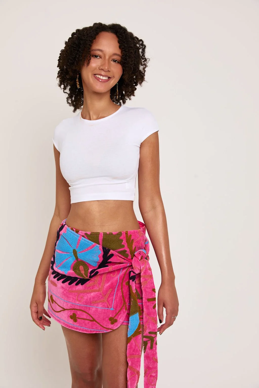A woman with curly hair smiling, wearing a white crop top and a colorful pink, blue, and green sarong tied around her waist, standing against a plain white background.