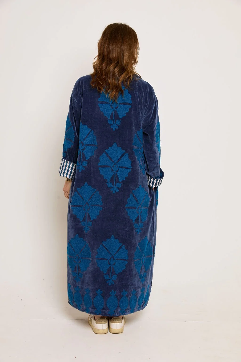 Back view of a woman wearing a long blue velvet coat with a large floral pattern, striped shirt cuffs, and beige sneakers
