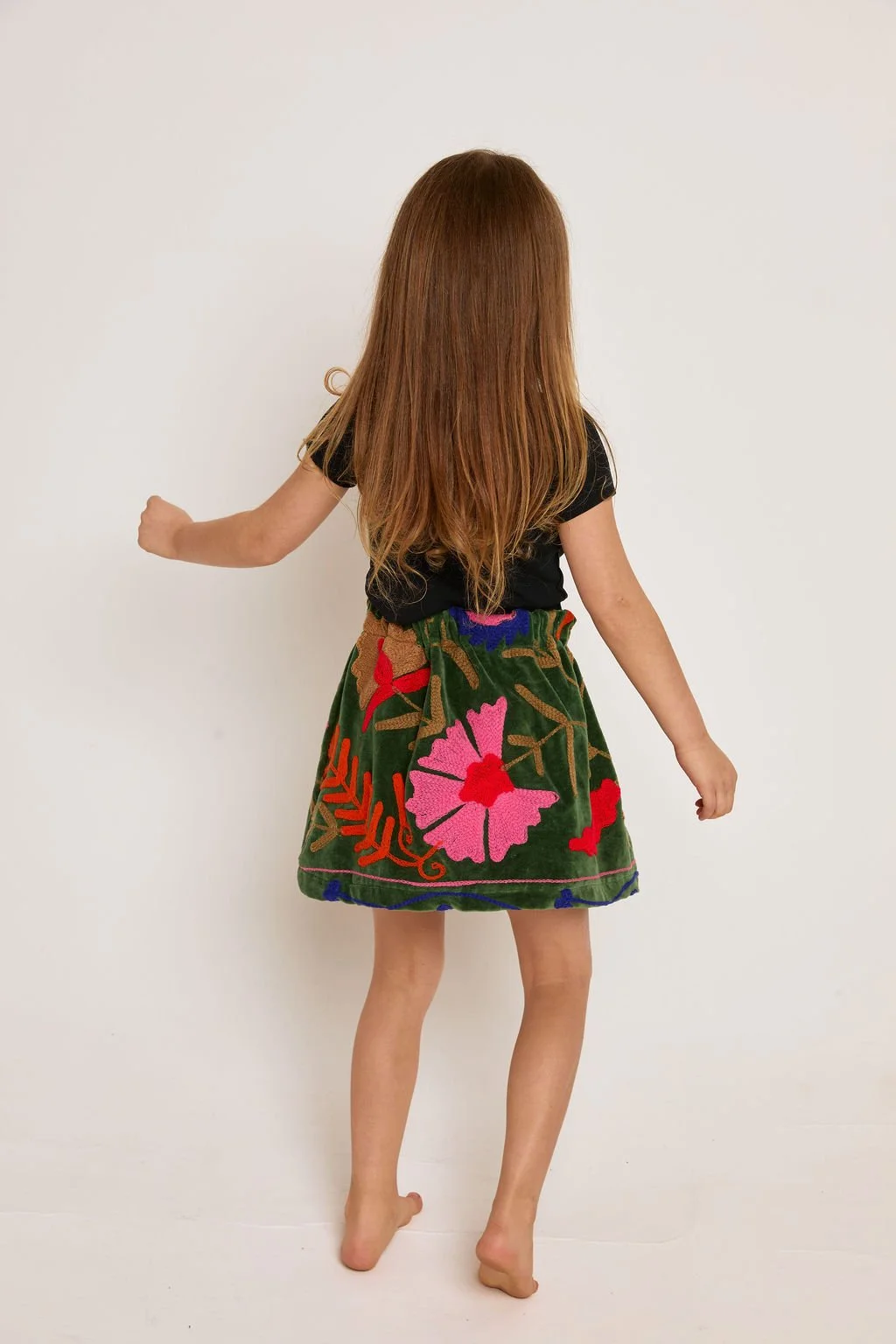 A young girl with long red hair, wearing a black top and a colorful embroidered skirt, facing away from the camera.